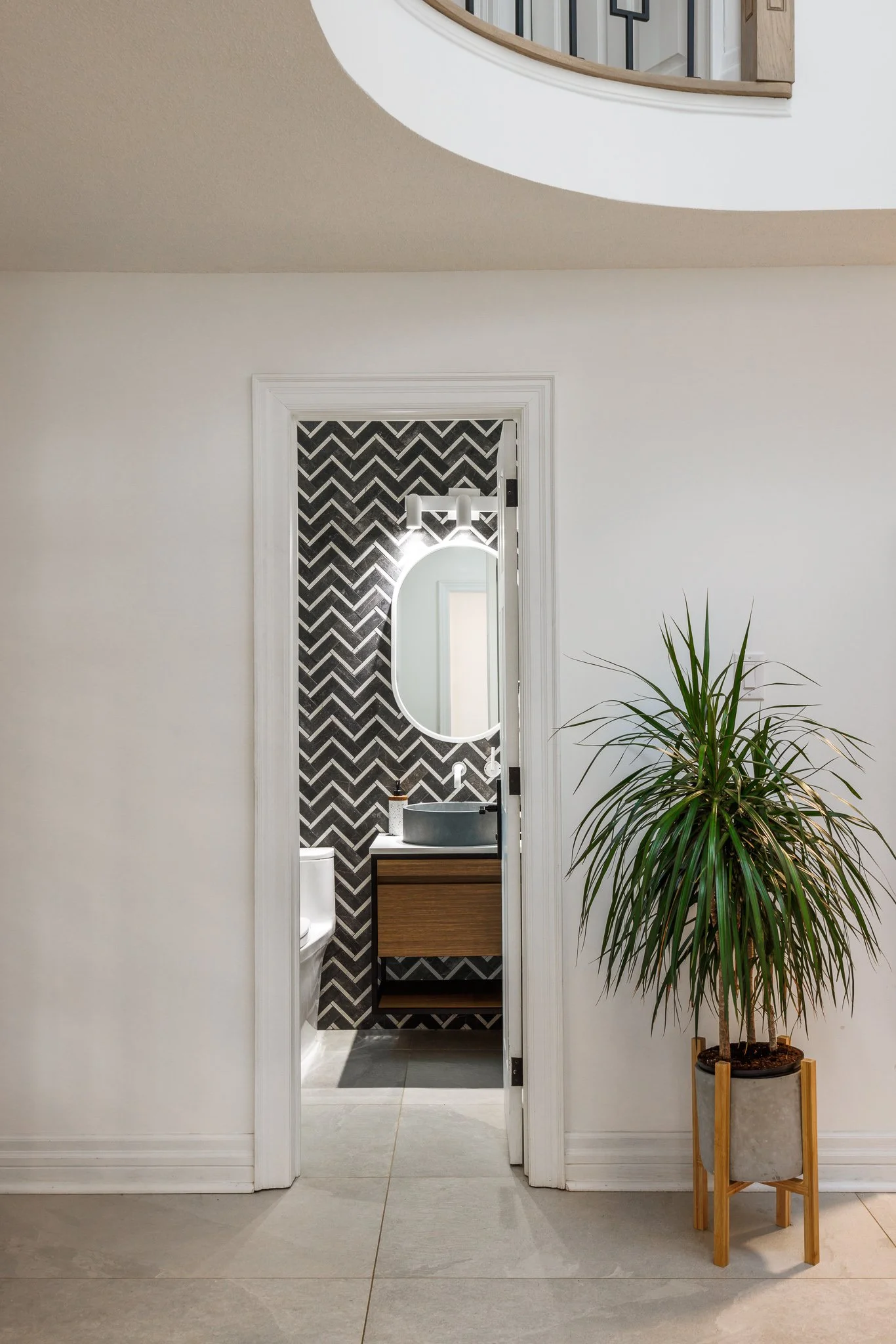 Interior view of a bathroom seen through a doorway, with a black and white zigzag tile wall, a round mirror, a stone vessel sink, and a wooden vanity, next to a potted plant.