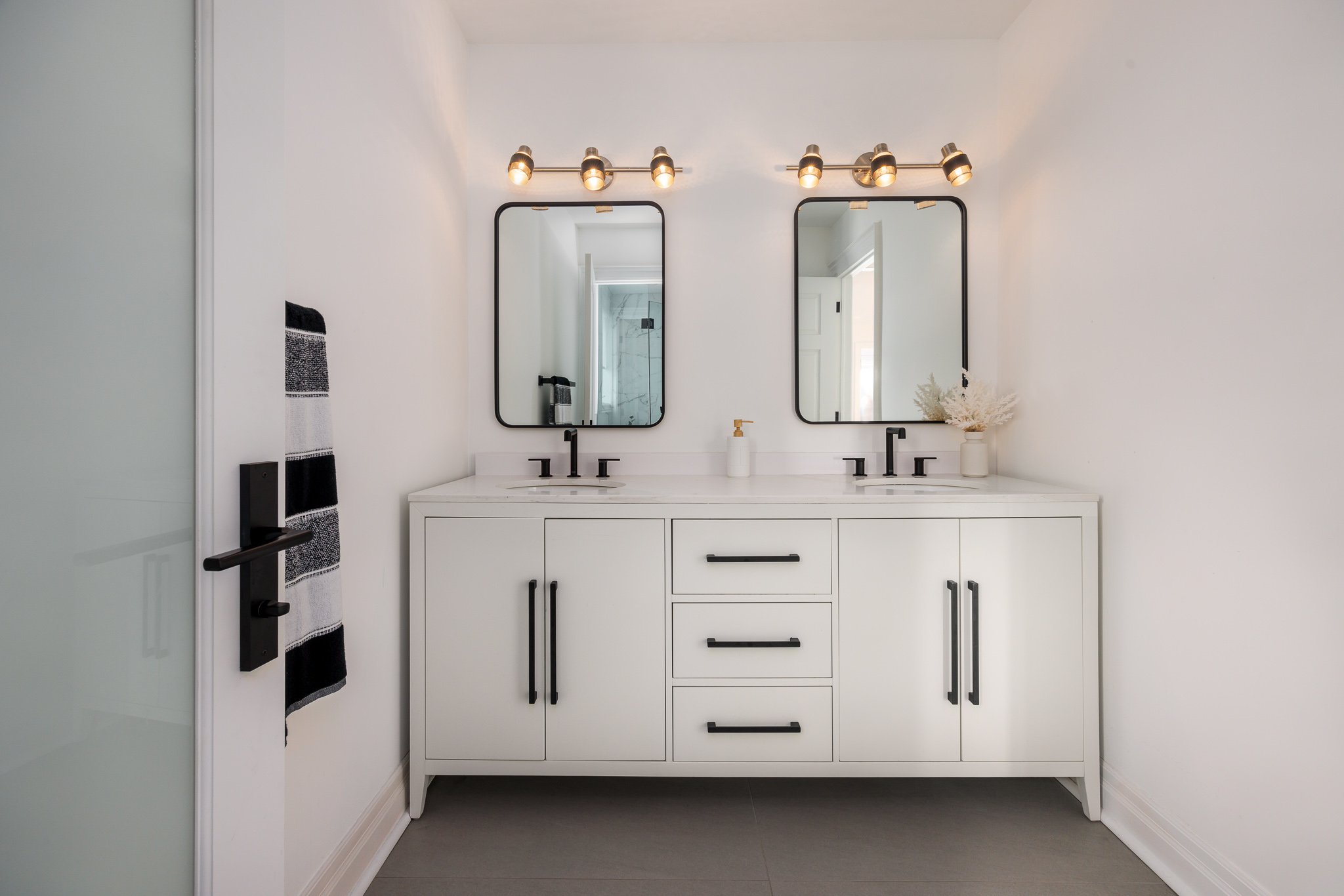Modern white bathroom vanity with dual sinks, black hardware, two mirrors, and black lighting fixtures.