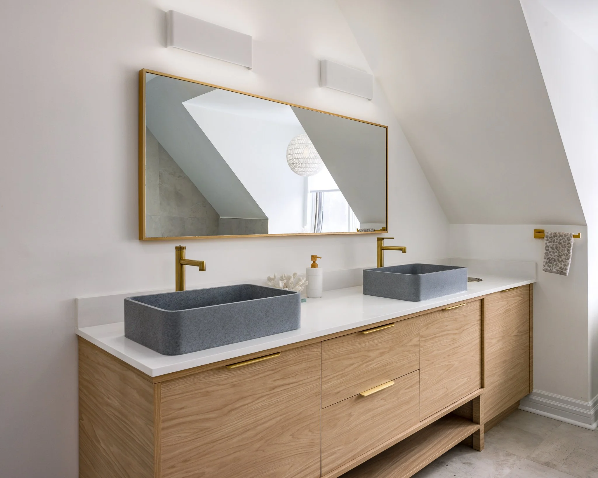 Modern bathroom vanity with twin grey stone vessel sinks, gold faucets, a large framed mirror, and a white countertop in a minimalist style.