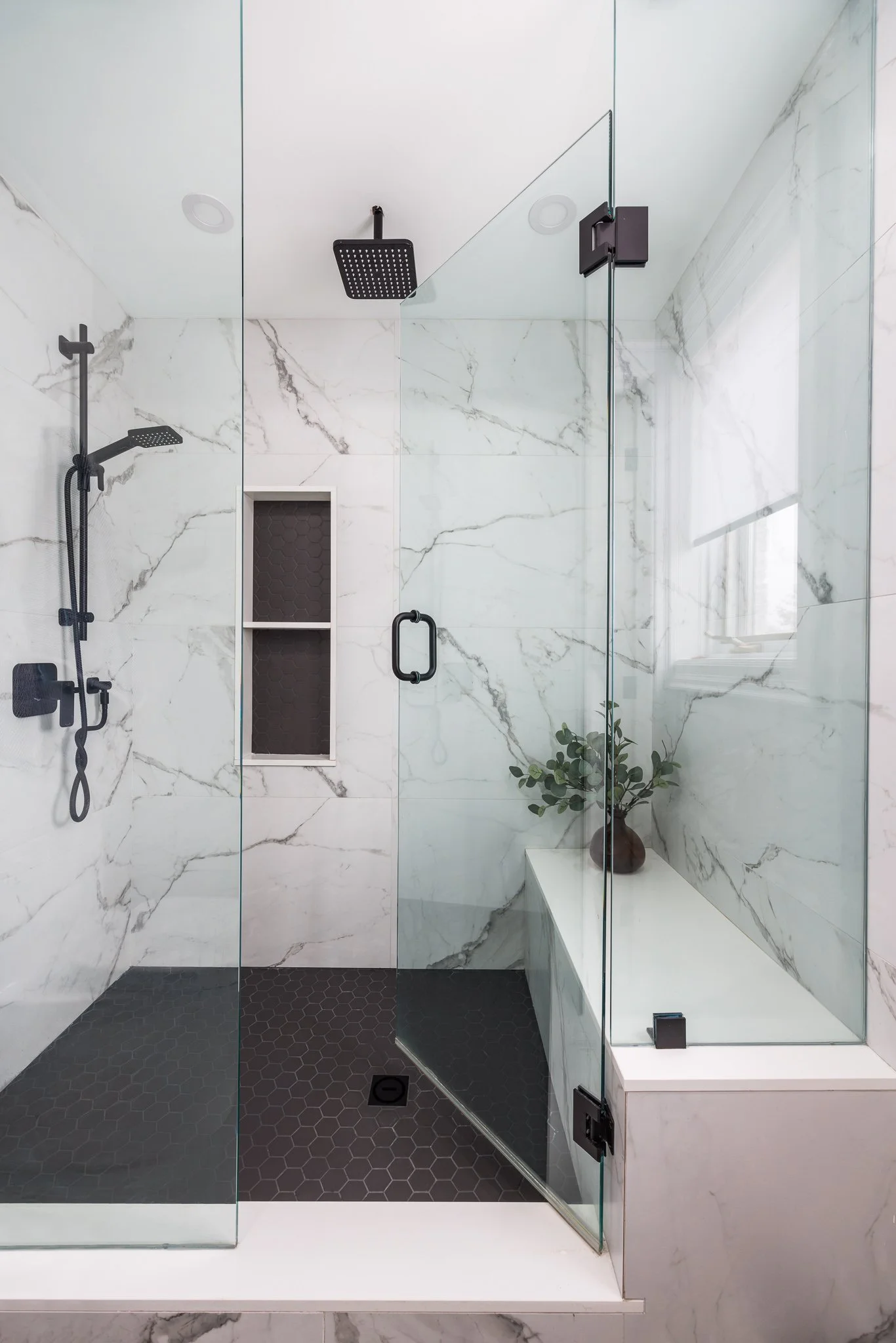Modern walk-in shower with white marble walls, black fixtures, and a glass door, featuring a built-in niche and a small bench with a potted plant.