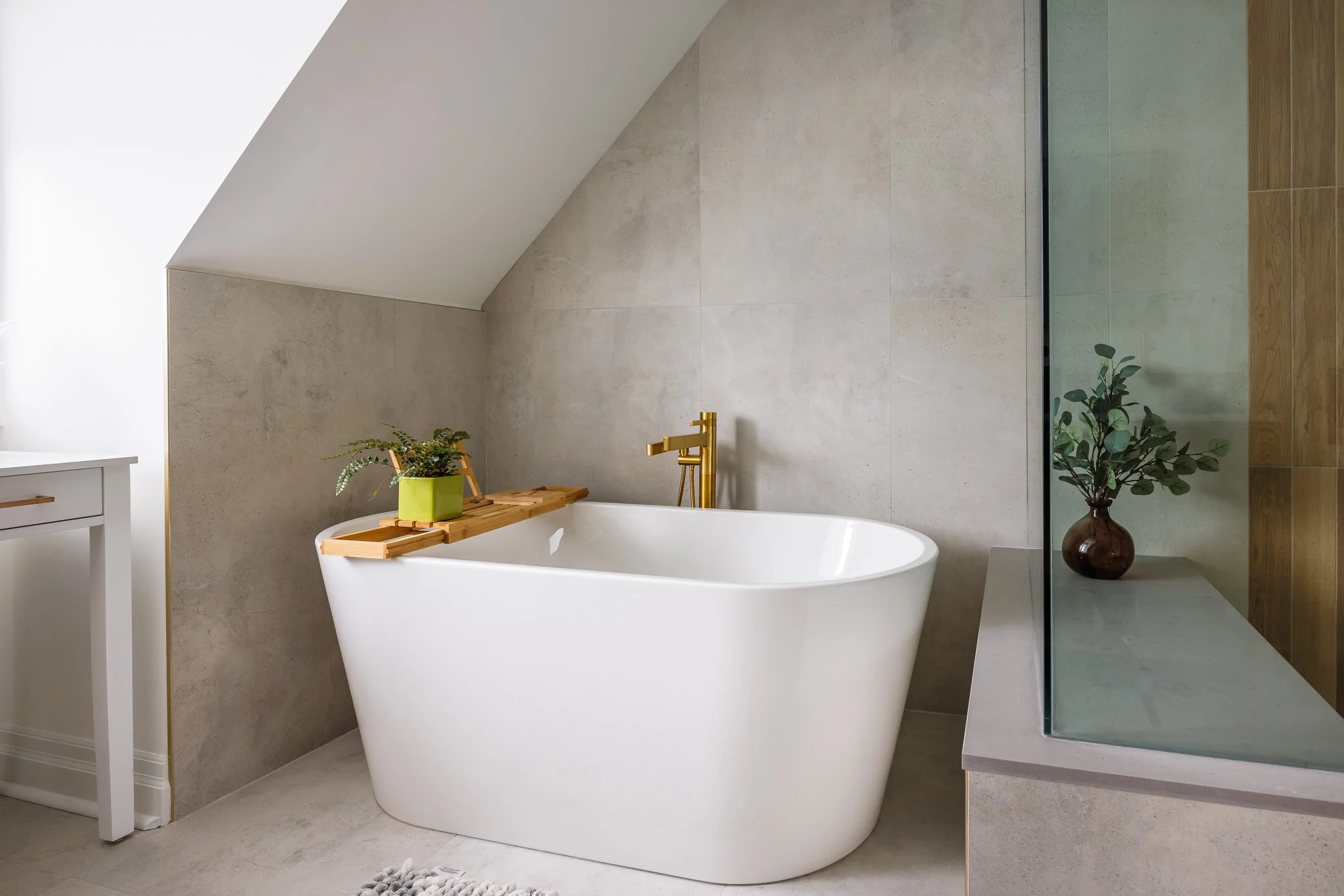 Modern bathroom with a white freestanding bathtub, gold faucet, and a small wooden tray with a green plant. There's a frosted glass shower enclosure with a potted plant inside, and neutral-colored tiles on walls and floor.