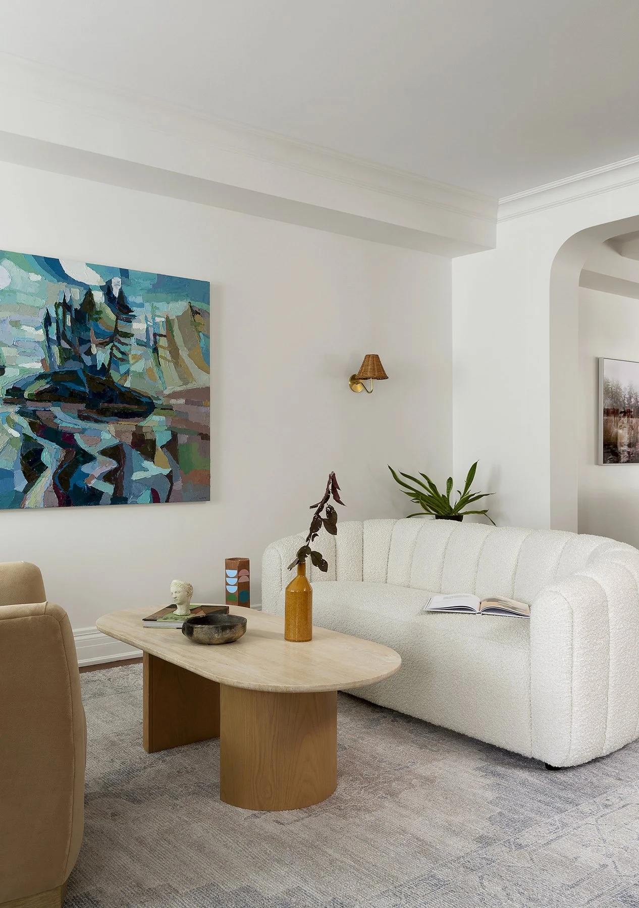 A living room with a white textured sofa, a light wood oval coffee table decorated with a tall yellow vase with dark branches, a small sculpture, and a decorative bowl. A large abstract painting with shades of blue, green, and purple hangs on the whi