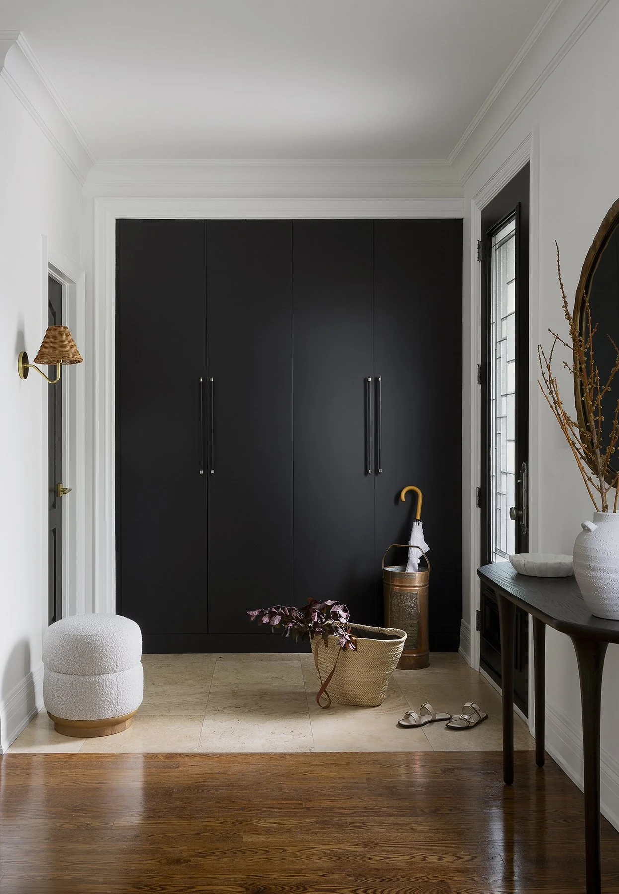 Interior entryway with black cabinetry, a white wall, a small white ottoman, a wicker basket with a plant, a pair of sandals, a side table with a white vase and dried branches, and an umbrella stand.