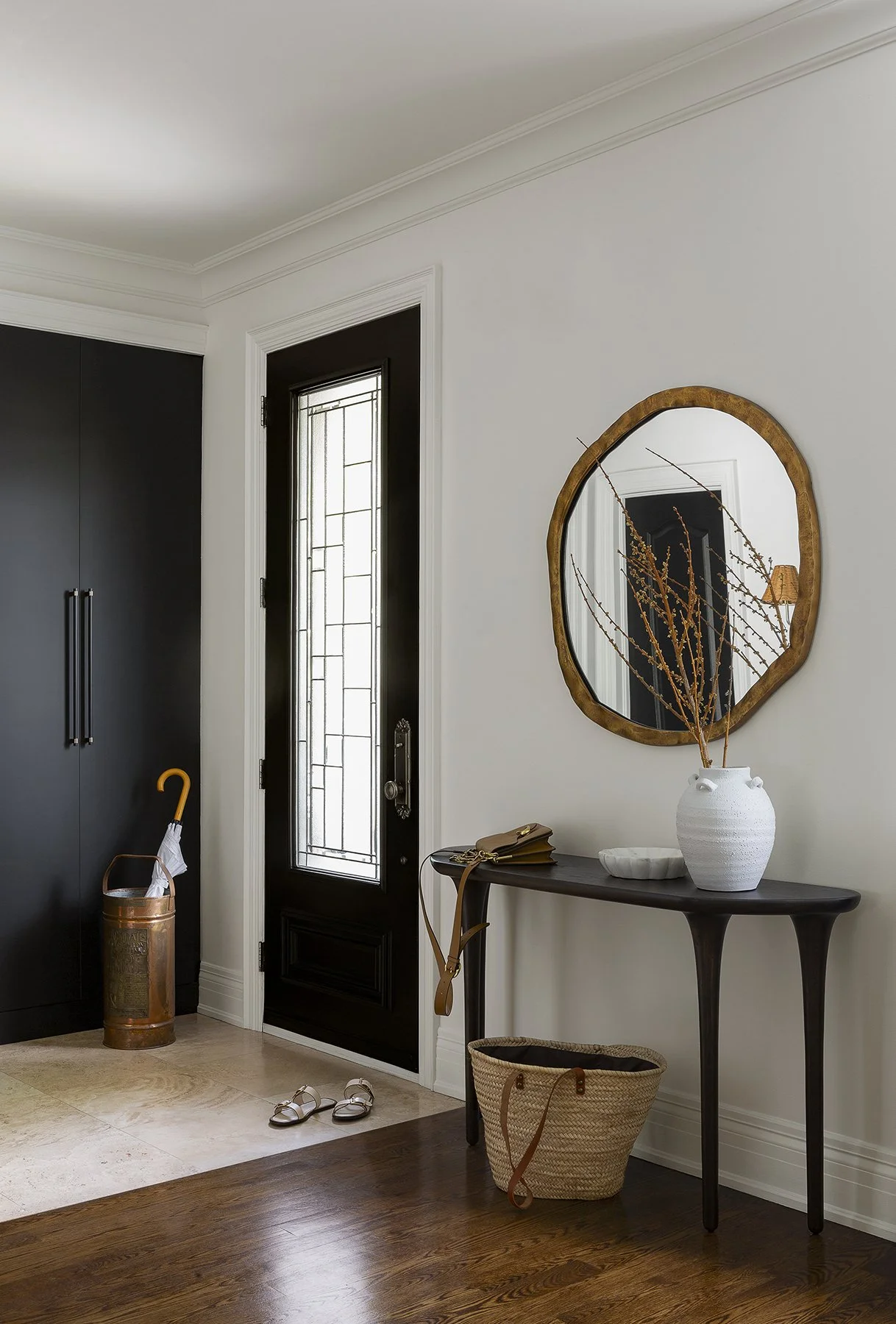 Interior entryway with black front door, white walls, a dark wood table with a white vase and dried branches, a mirror with a wooden frame, and a pair of sandals on a tiled floor.