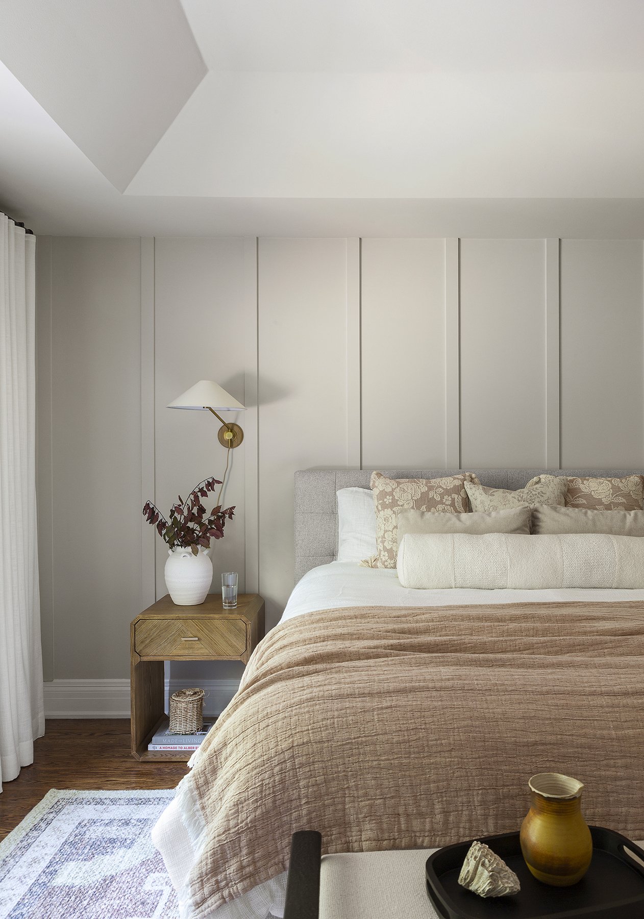 A cozy bedroom with a bed featuring beige and cream pillows, a wooden nightstand with a white vase containing reddish-brown leaves, a glass of water, and a brass wall-mounted lamp. There is a beige throw blanket on the bed, a patterned rug on the woo