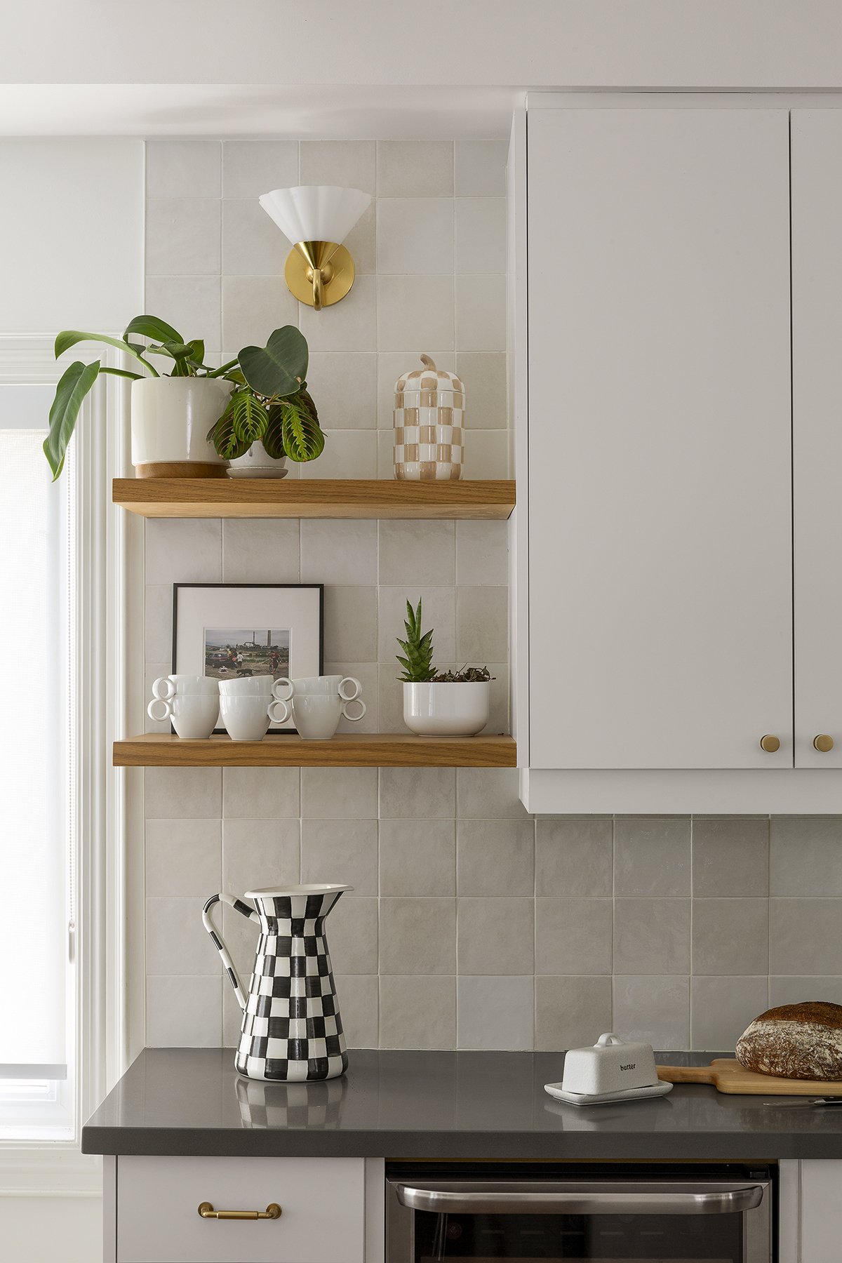 Kitchen shelf with potted plant, ceramic decor, framed photo, and white cups, against tiled wall with wall light, with a black-and-white checkered pitcher on the counter.