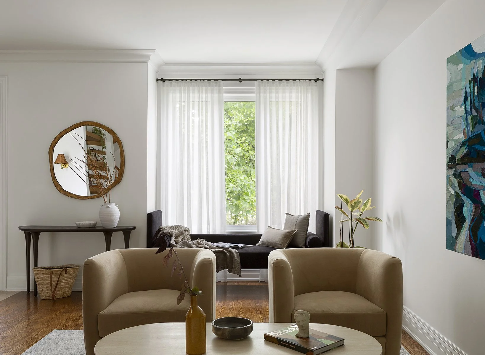 Bright living room with beige chairs, a black window seat with pillows, a side table with a white vase and decorative items, a mirror, artwork, and plants, with natural light coming through sheer curtains.