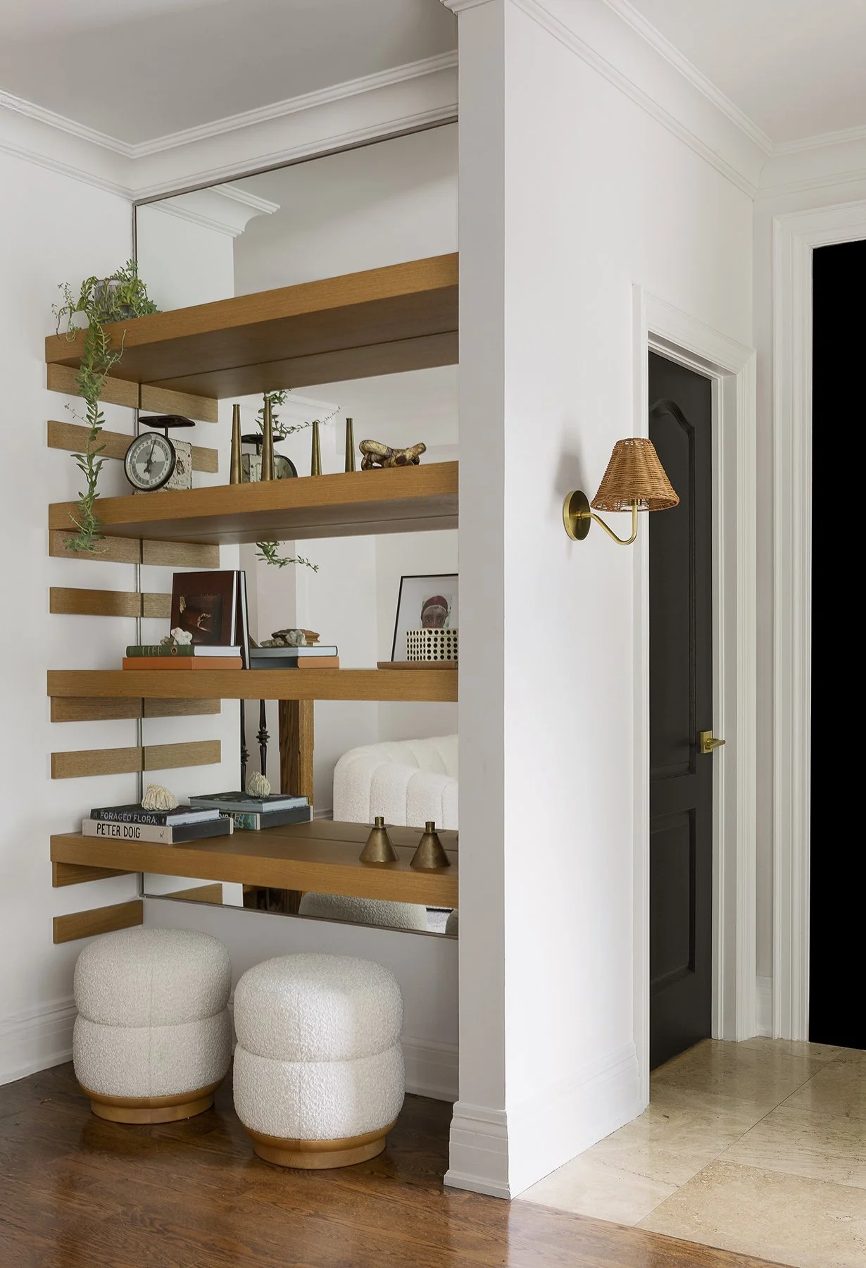 Part of a modern living room, showing a white wall with a brass wall sconce, a black door, wooden and glass shelves with decorative items, books, and small sculptures, and two white textured ottomans on a wooden floor.