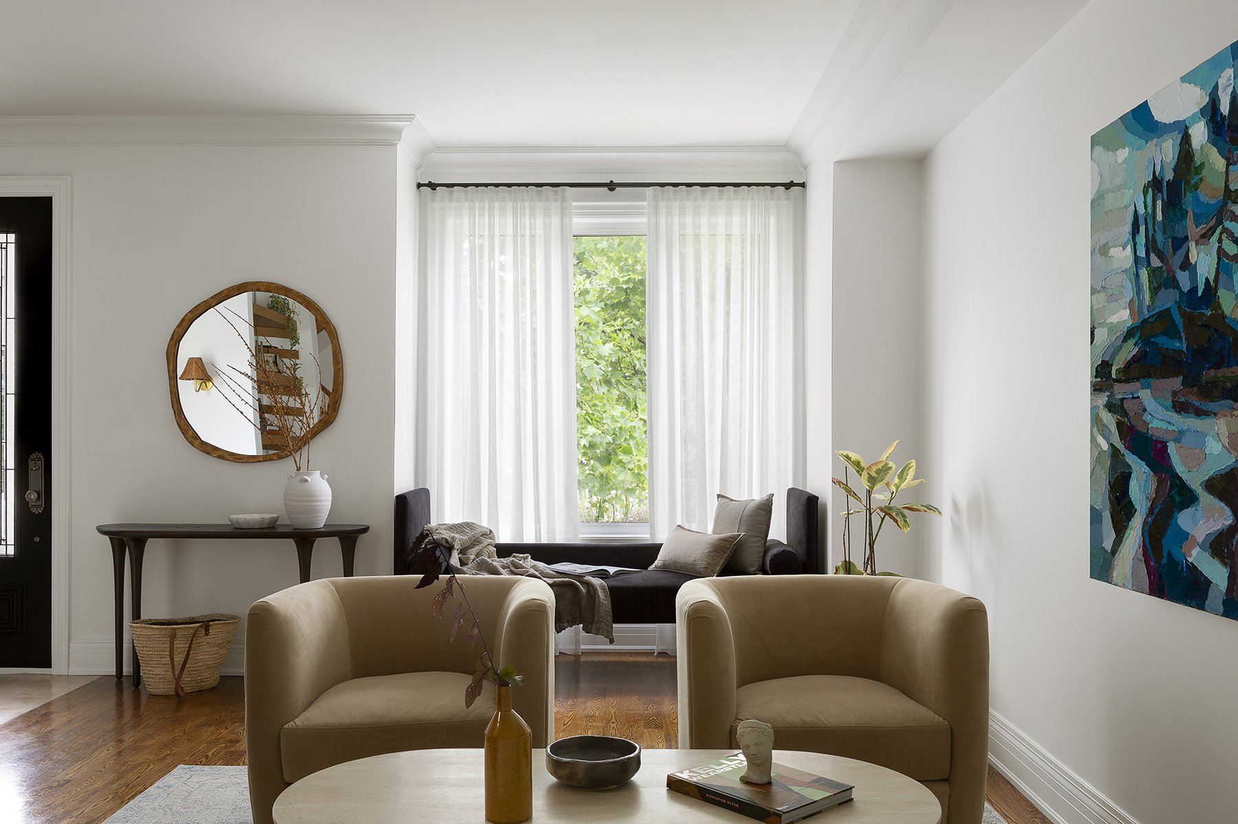 Living room with two beige armchairs, a coffee table with decorative items, a window with sheer white curtains, a black bench with cushions, and wall art.