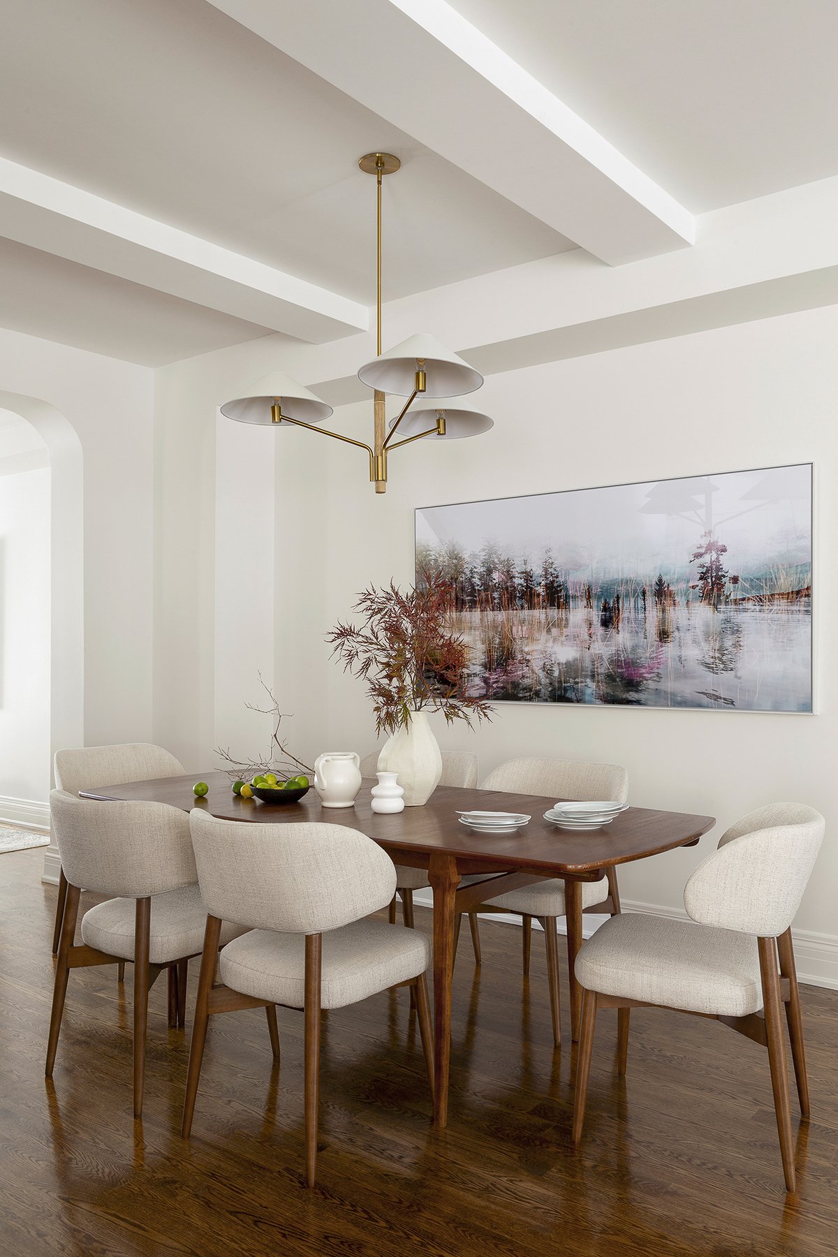 A dining room with a wooden table, six beige upholstered chairs, a modern gold and white chandelier, a large landscape art piece on the wall, and a vase with branches as centerpiece.