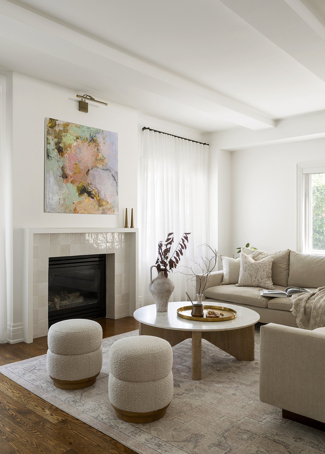 Bright living room with white walls, fireplace, abstract painting, cream sofa with pillows, round coffee table, two round ottomans, and decorative vases.