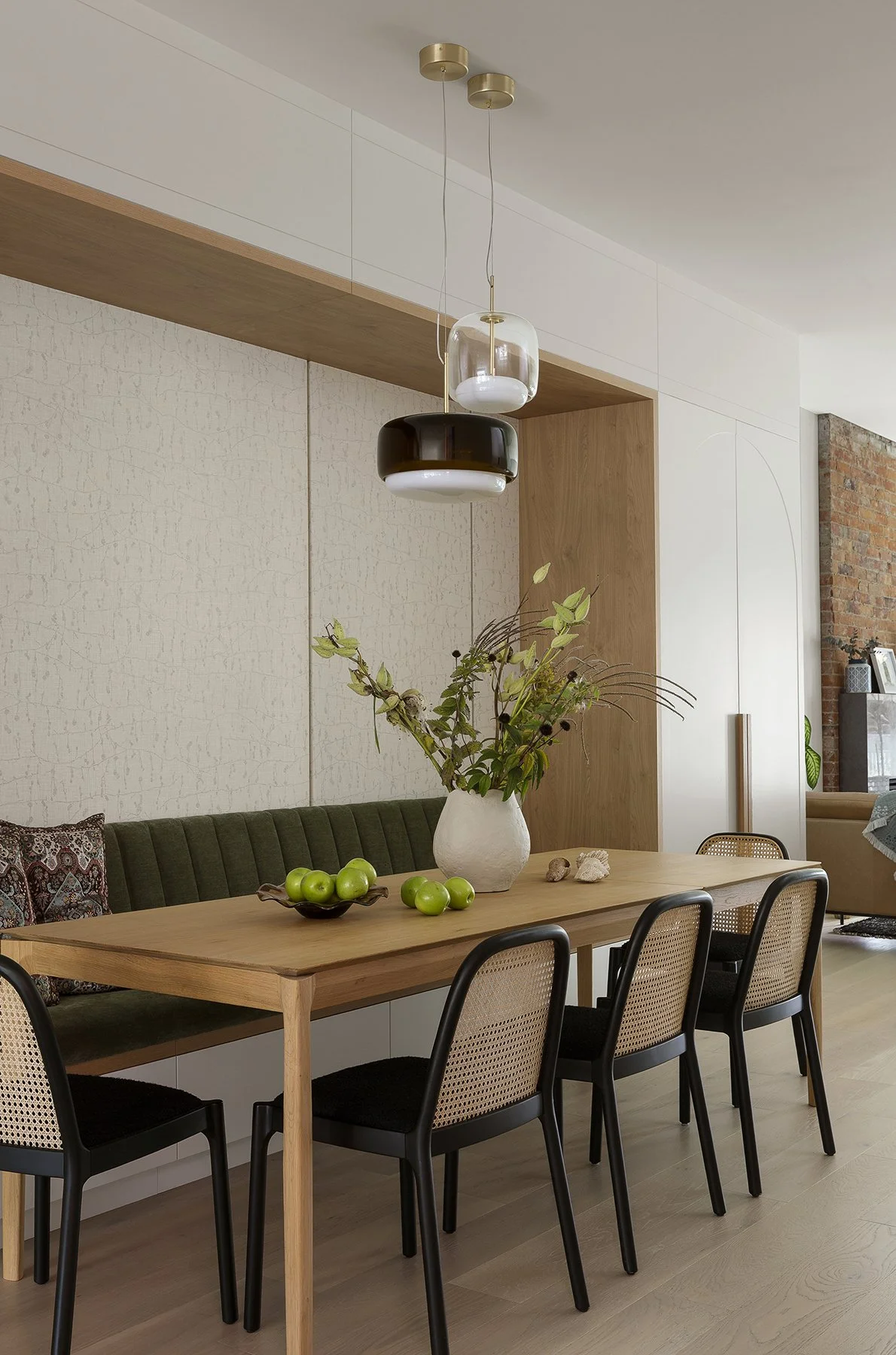 A modern dining room with a wooden table, black chairs, and a green velvet bench. A white vase with green foliage and green apples decorate the table. Hanging above are three pendant lights in different sizes and colors.