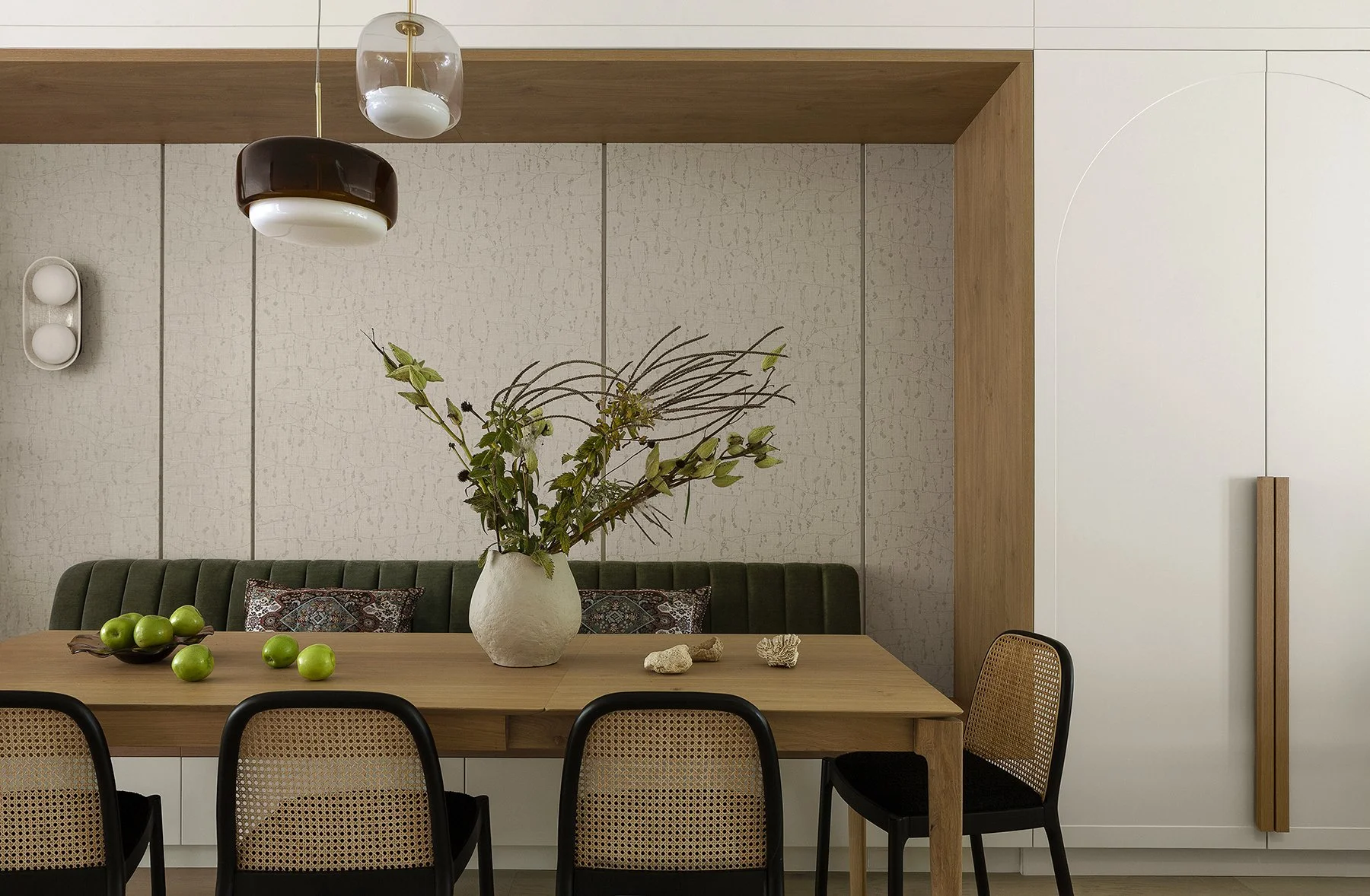 Dining room with a wooden table, green cushioned bench, and black chairs. A large white vase with green foliage and branches, along with a few green apples and seashells, are on the table. White and beige textured walls with subtle vertical lines, a 