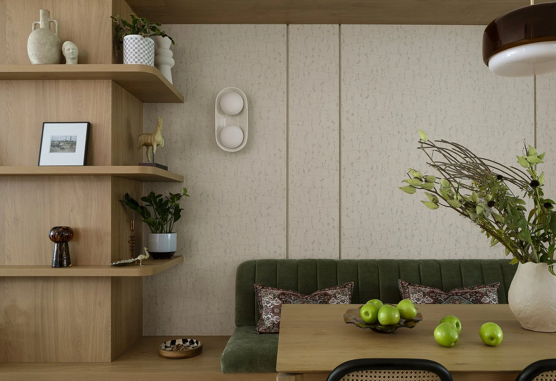 A living room corner with a wooden shelf filled with decorative items and plants, a green upholstered bench, a wooden table with green apples and a large vase with branches, and a beige textured wall with wall decor.