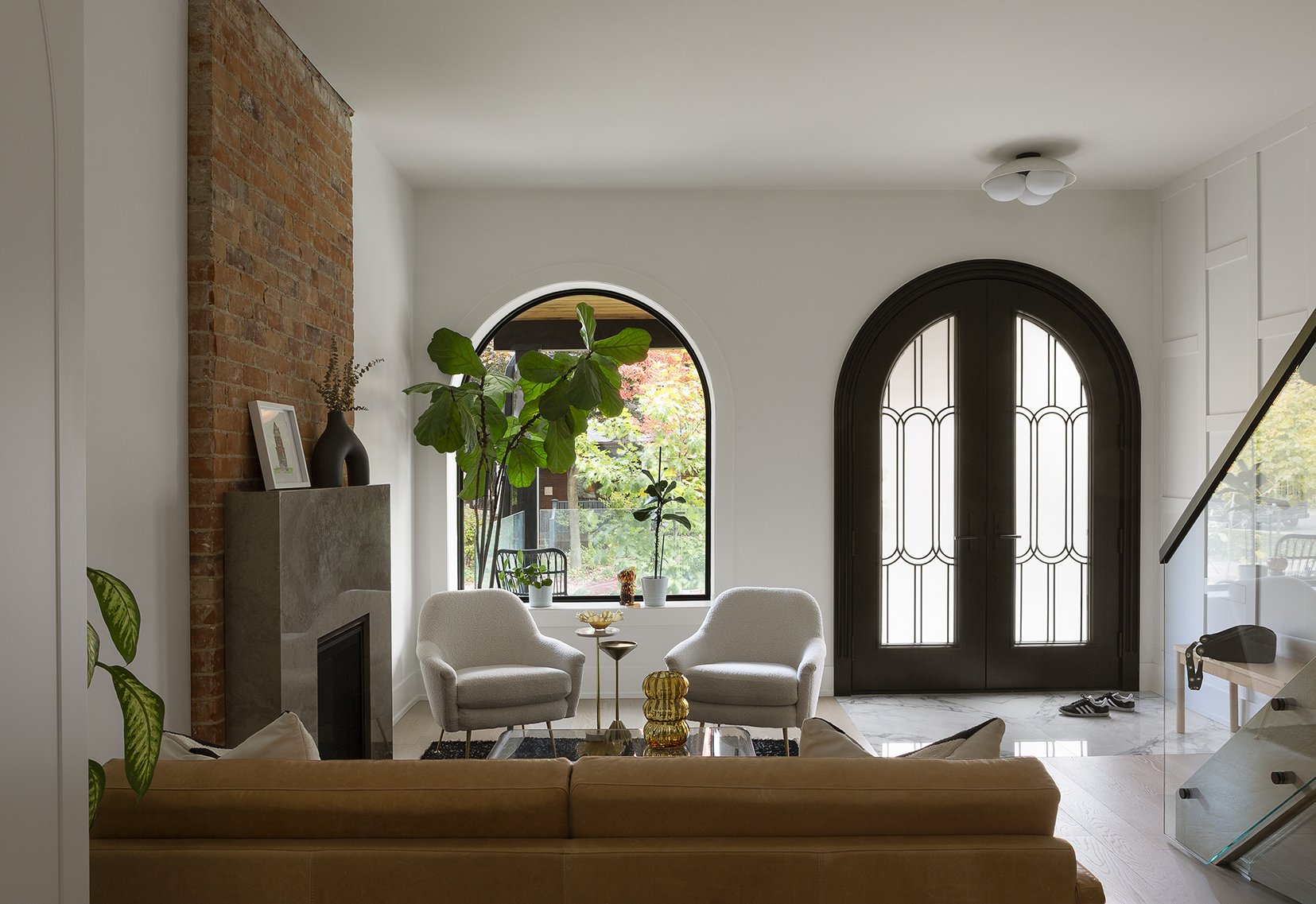 Living room with white walls, a brick fireplace, large arched window, dark arched front door with glass panels, two white armchairs, a glass coffee table with decorative objects, plants near the window, and a beige sofa in the foreground.