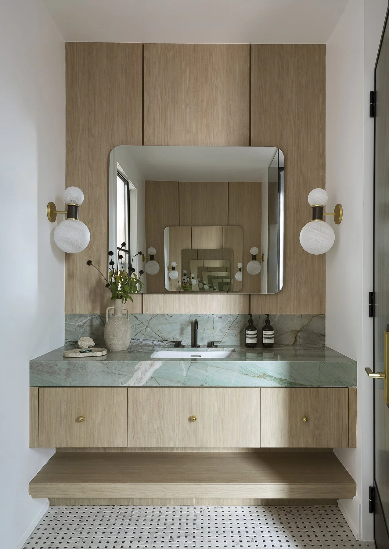 Modern bathroom vanity with a green marble countertop, a rectangular undermount sink, a large mirror reflecting an intricate pattern, wall-mounted white globe lights on both sides, a vase with dried flowers, and two black soap dispensers.