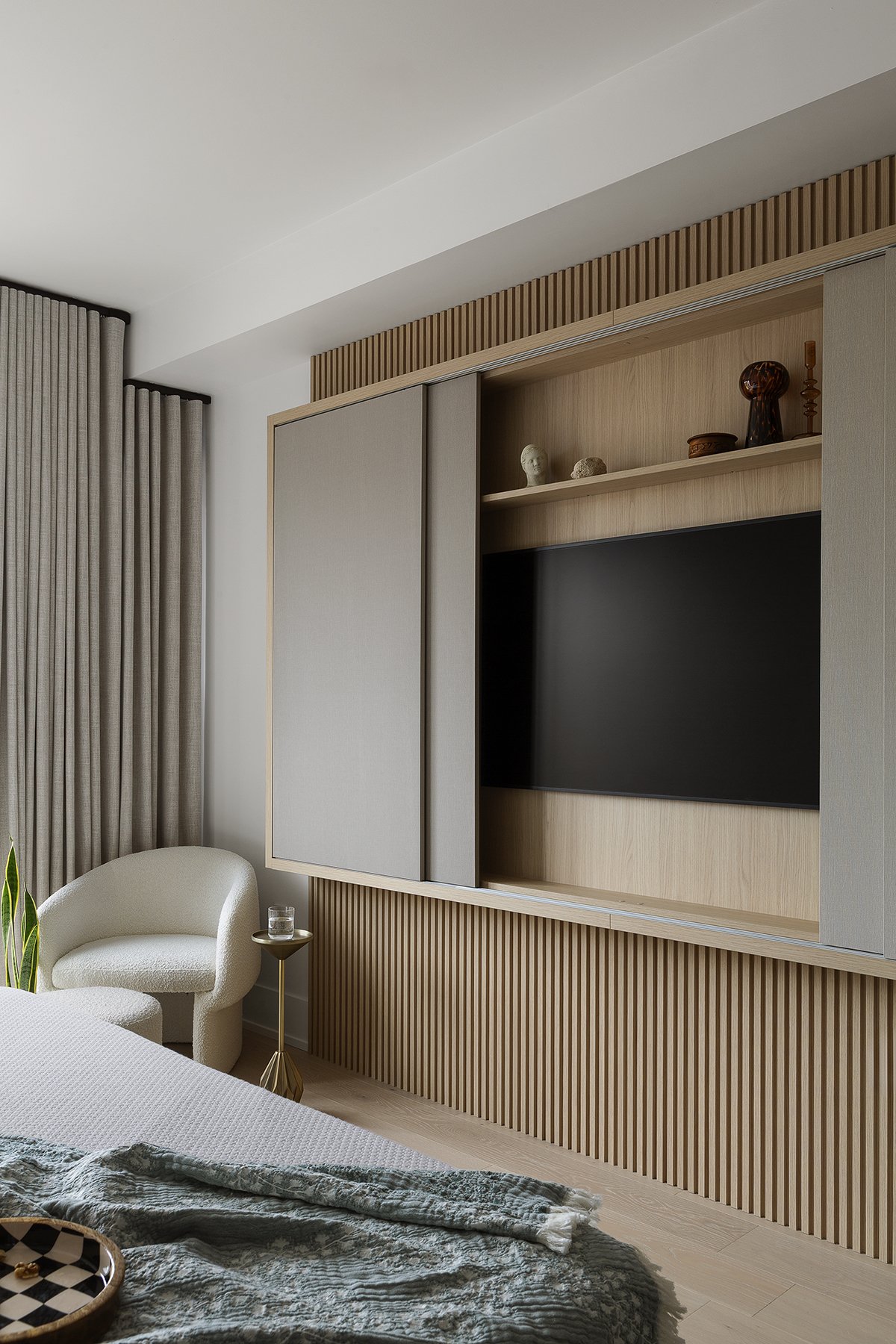 Modern bedroom with a wall-mounted TV, built-in wooden shelves displaying decorative items, beige curtains, an armchair with a side table and glass, and a bed with textured bedding.