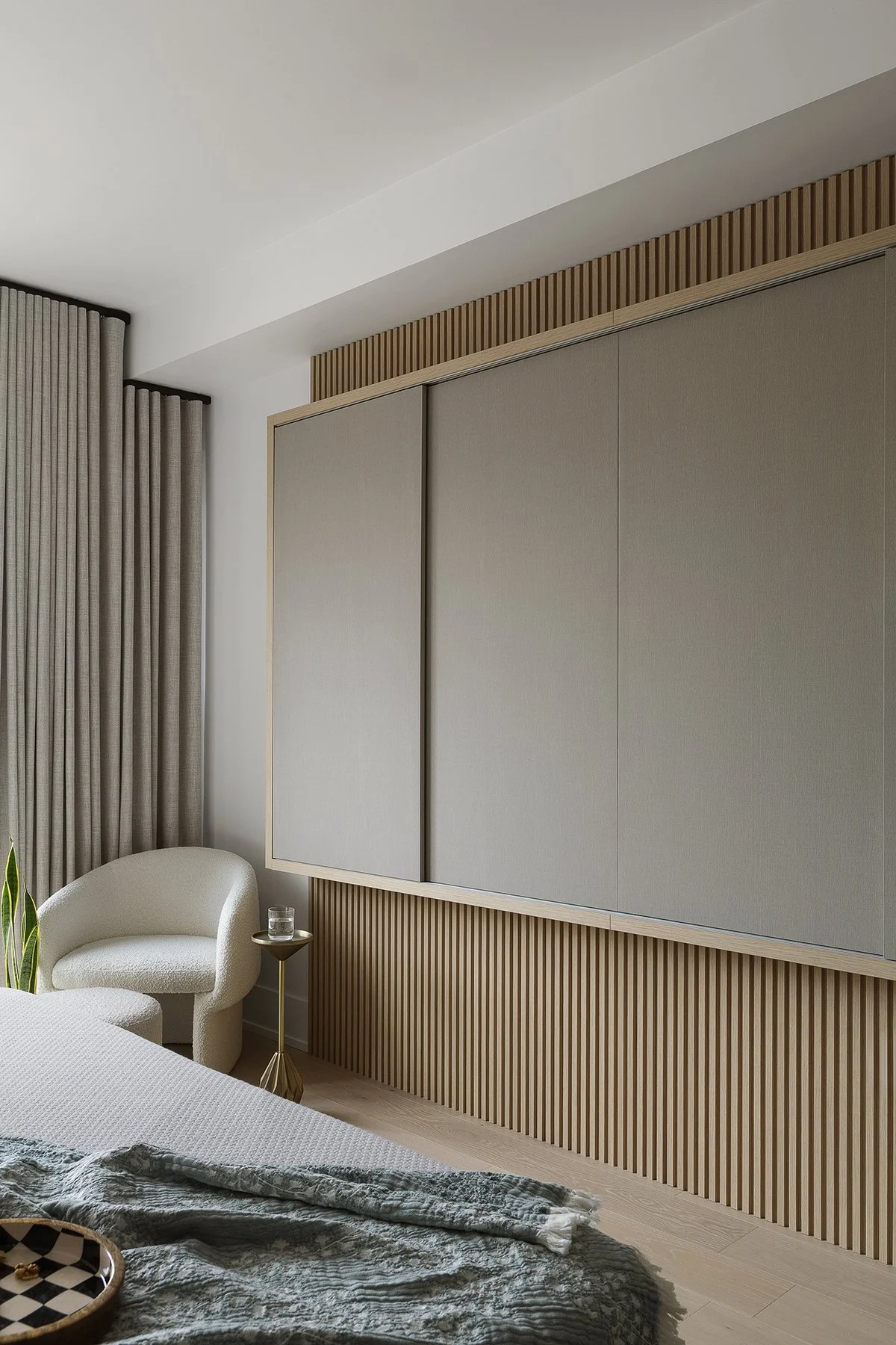 Modern bedroom with beige sliding closet doors, a white armchair, side table with glass of water, curtains, a bed with a textured blanket, and natural wooden elements.