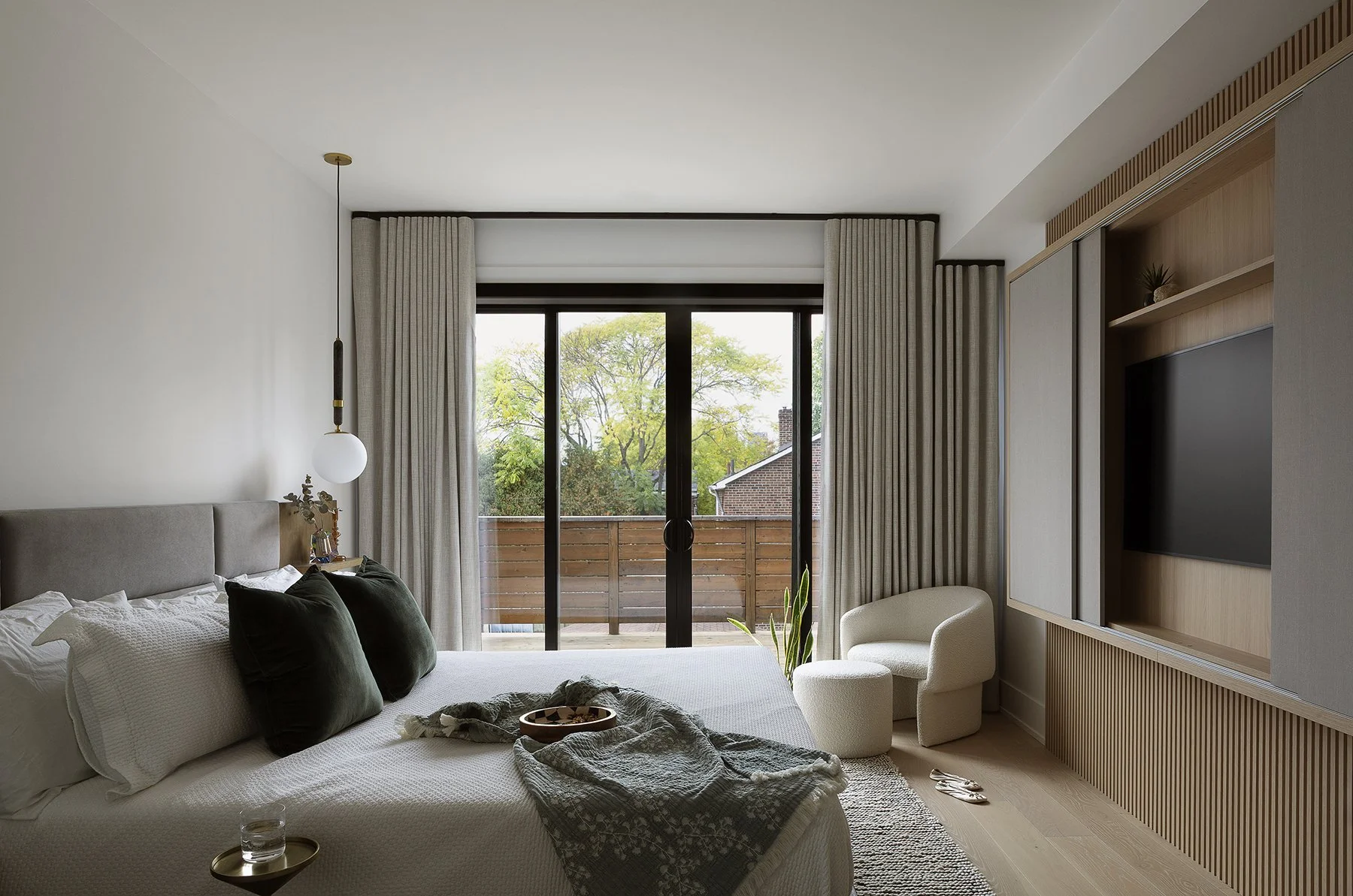 Modern bedroom with large sliding glass door leading to balcony, beige curtains, white bed with dark pillows, side table with water glass, armchair, and wooden accent wall with TV.