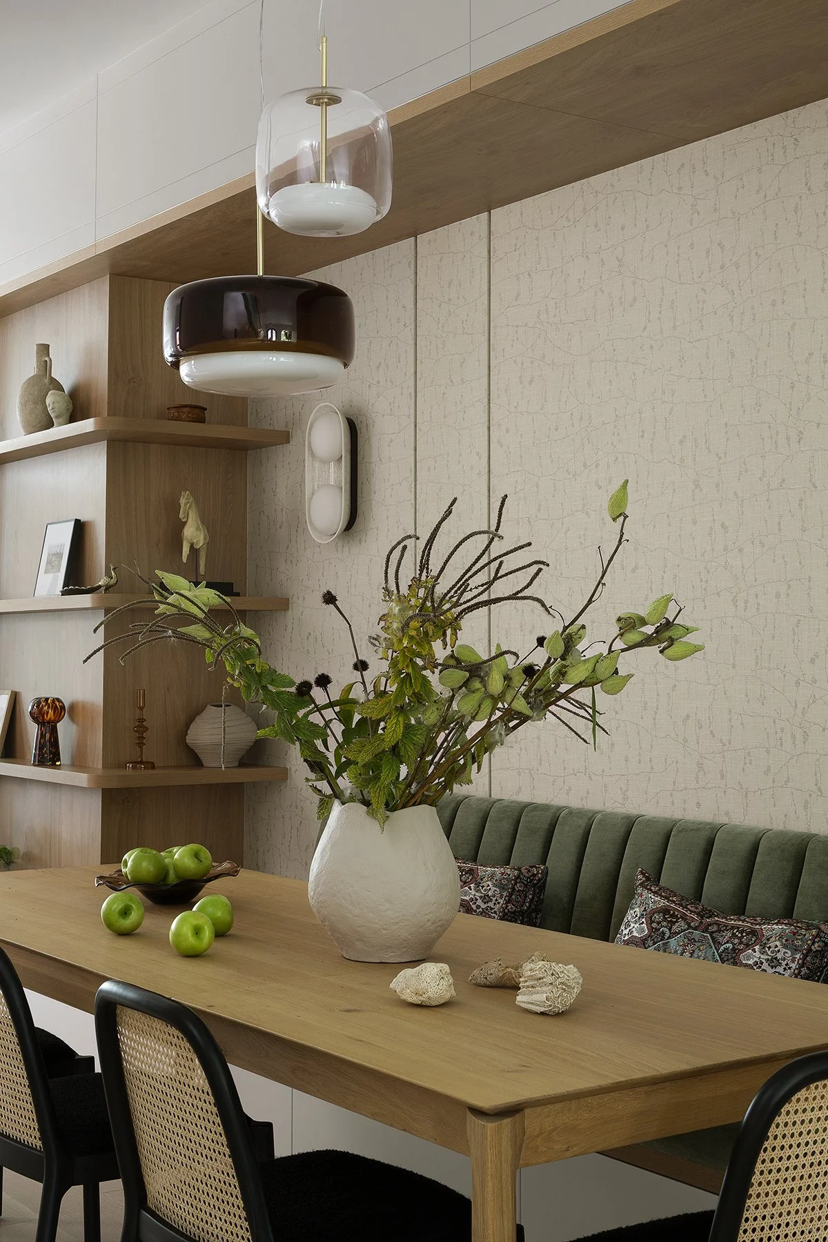 A modern dining room with a wooden table, green cushioned bench, black chairs, and a large white vase with greenery and branches. Shelves with decorative items and hanging light fixtures are visible in the background.
