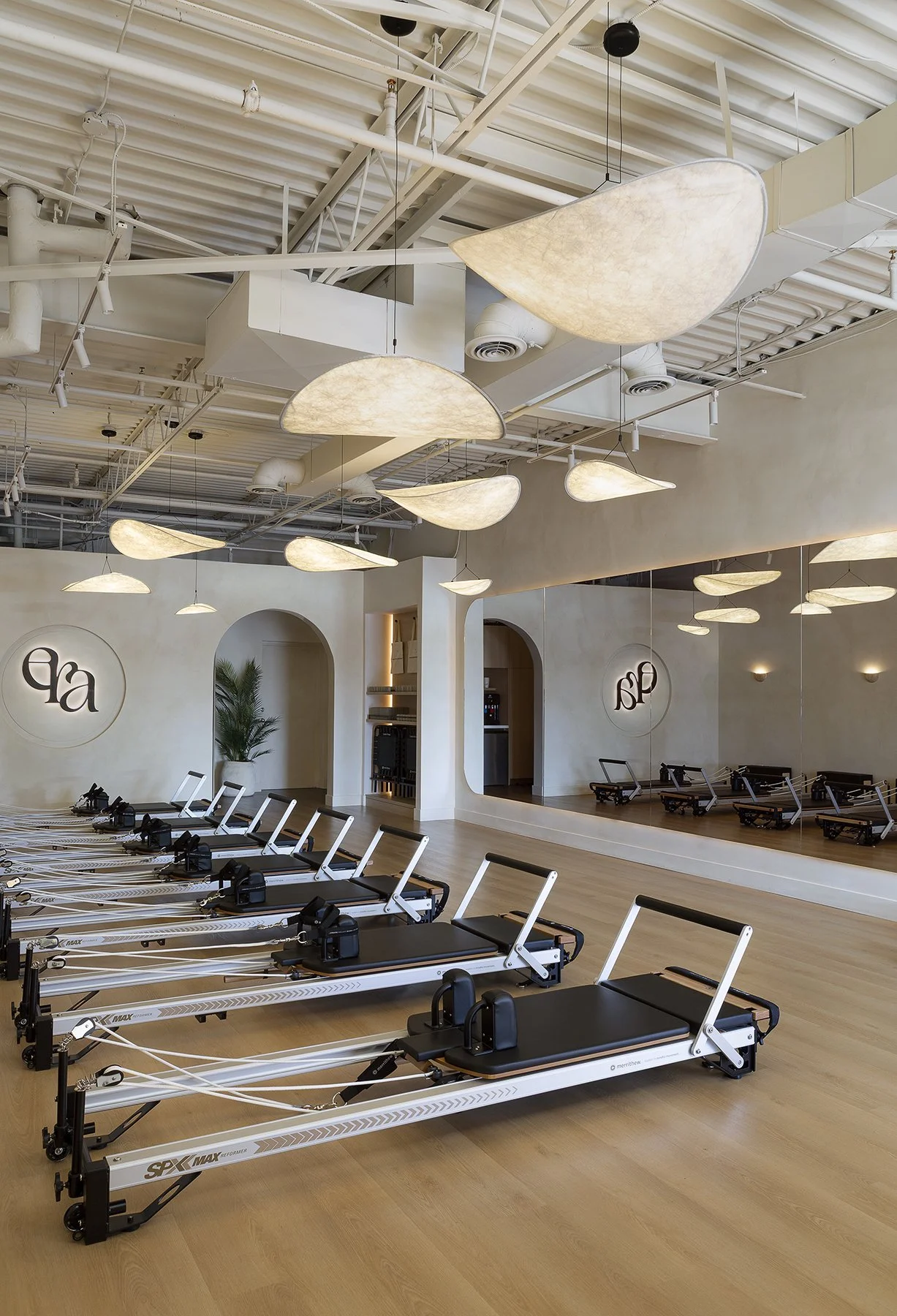Fitness studio with multiple reformer Pilates machines arranged in a row on a wooden floor, large ceiling lights, and a wall mirror reflecting the room.