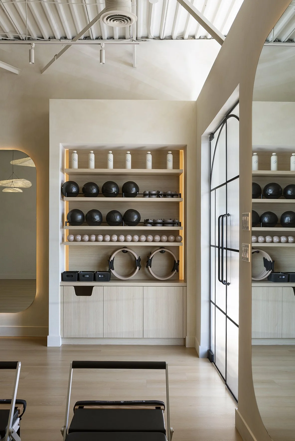 Interior of a fitness studio with shelves holding black and white exercise equipment, including kettlebells and headphones, finished in light wood with natural light coming through a nearby window.