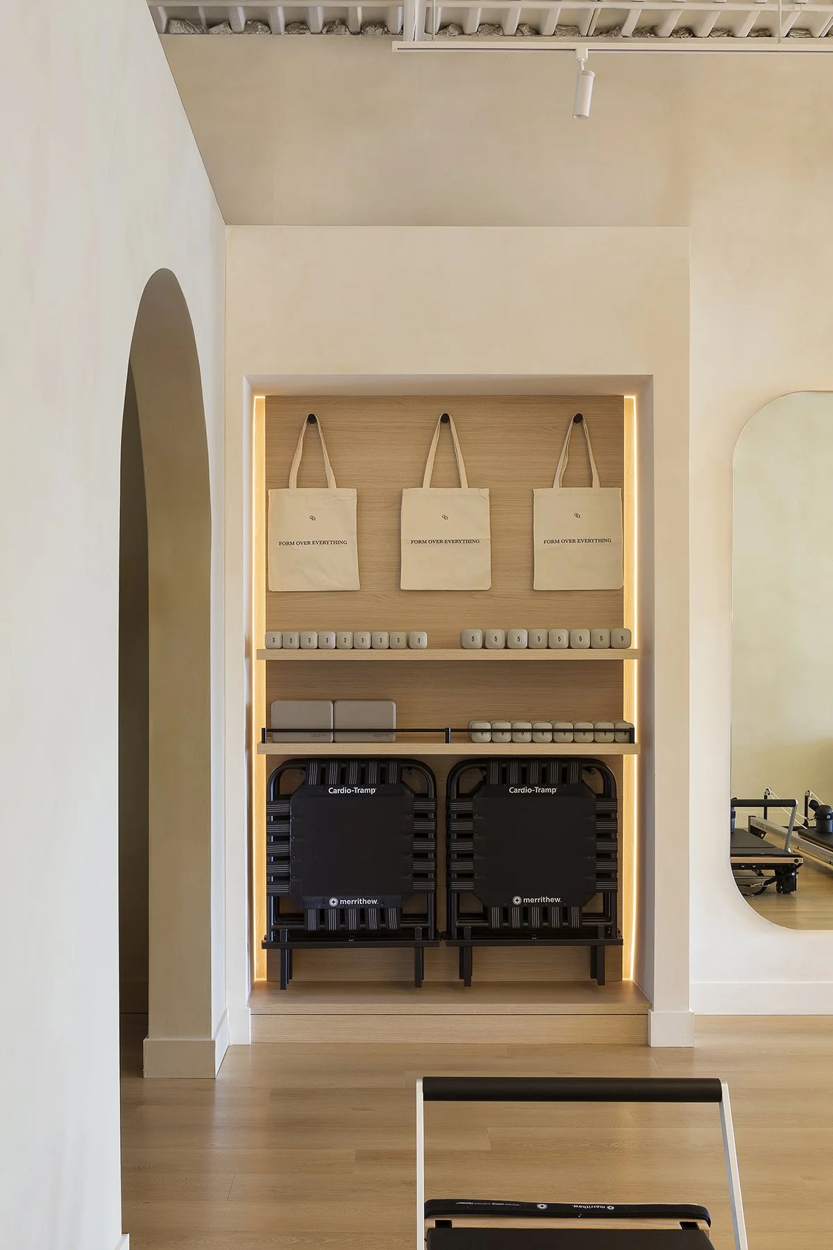 Workout studio organized with two Merrithew Cardio Tramps, storage shelves with tote bags labeled 'Form Over Everything', and a mirror on the right wall.