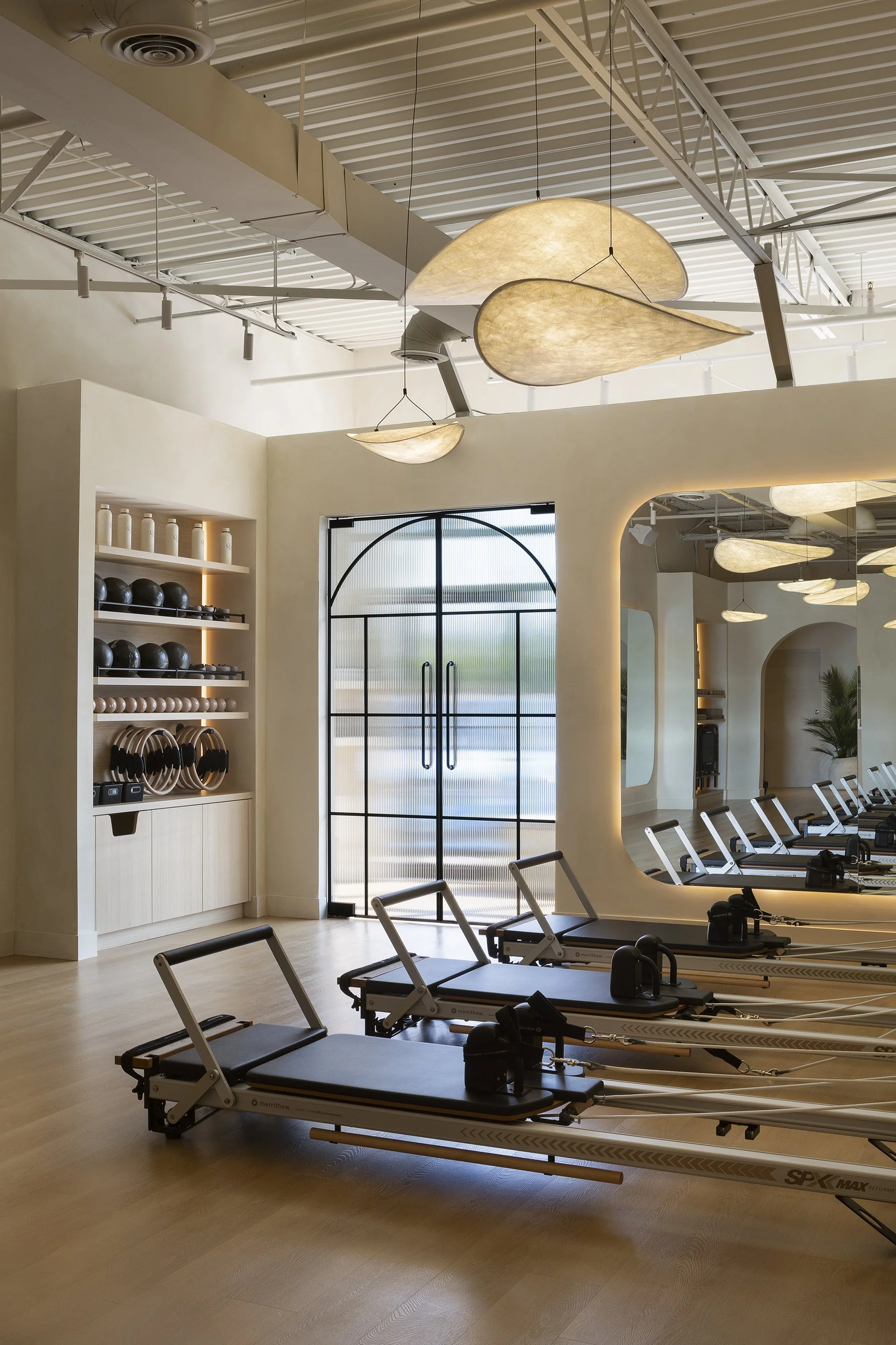 A modern gym with rowing machines, a shelf with kettlebells and workout accessories, and large decorative ceiling lights.