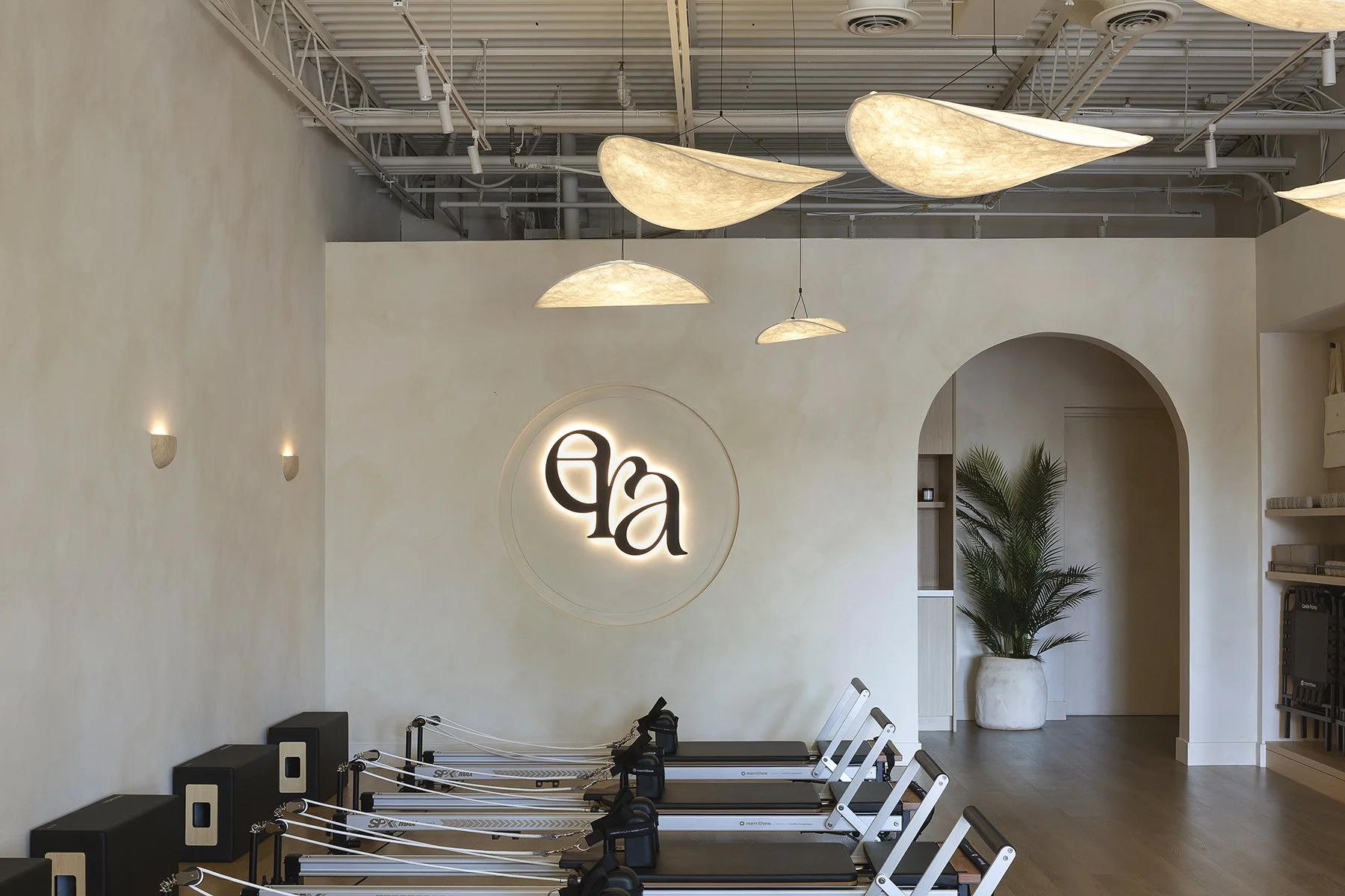 Interior of a fitness studio with light-colored walls, modern lighting fixtures on the ceiling, a large illuminated logo on the back wall, and several exercise machines lined up in the foreground near a potted plant.
