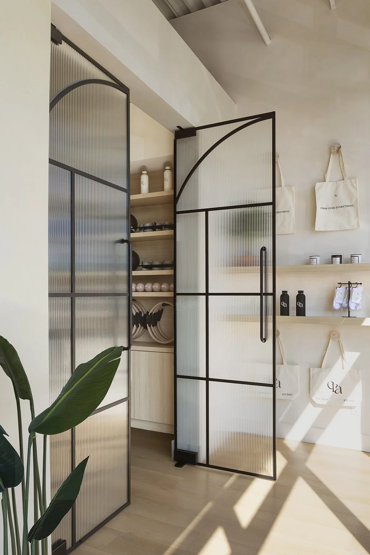 Interior space with a metal-framed glass door, open to reveal shelves with water bottles, bowls, and other items. There are white tote bags hanging on the wall and a wooden shelf with black bottles and small items. A large green plant is in the foreg
