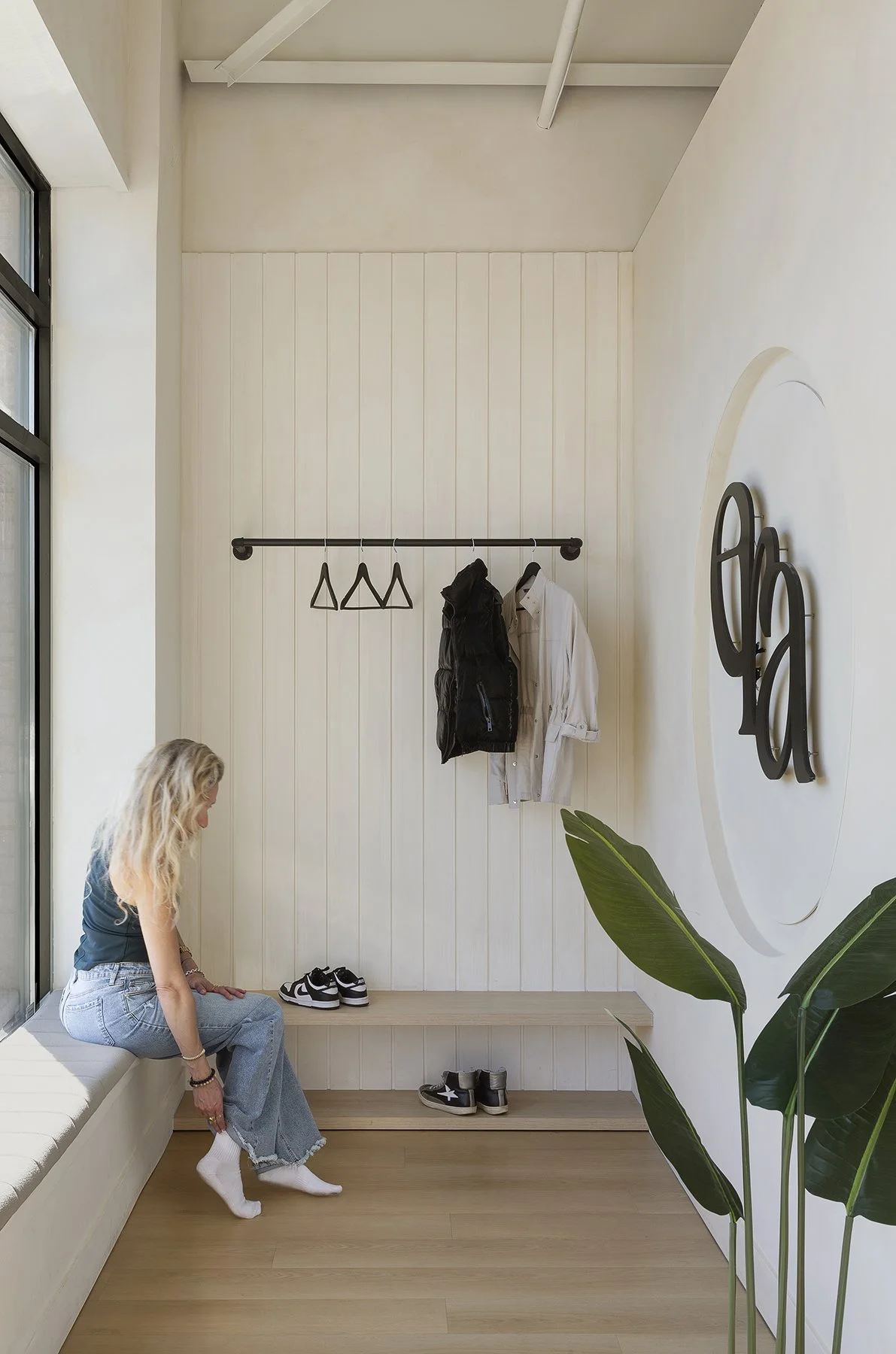 A woman sitting on a bench by a large window, with a modern interior and a wall-mounted coat rack holding a black vest and white shirt.