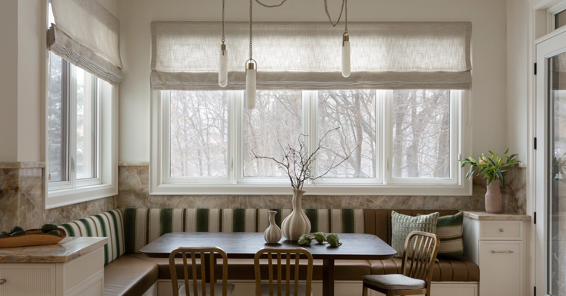 A cozy dining nook with large windows and beige Roman shades. A dark wooden table is centered, decorated with white vases and artichokes. The seating includes an L-shaped bench with striped cushions and two chairs, one with a green striped pillow. A 