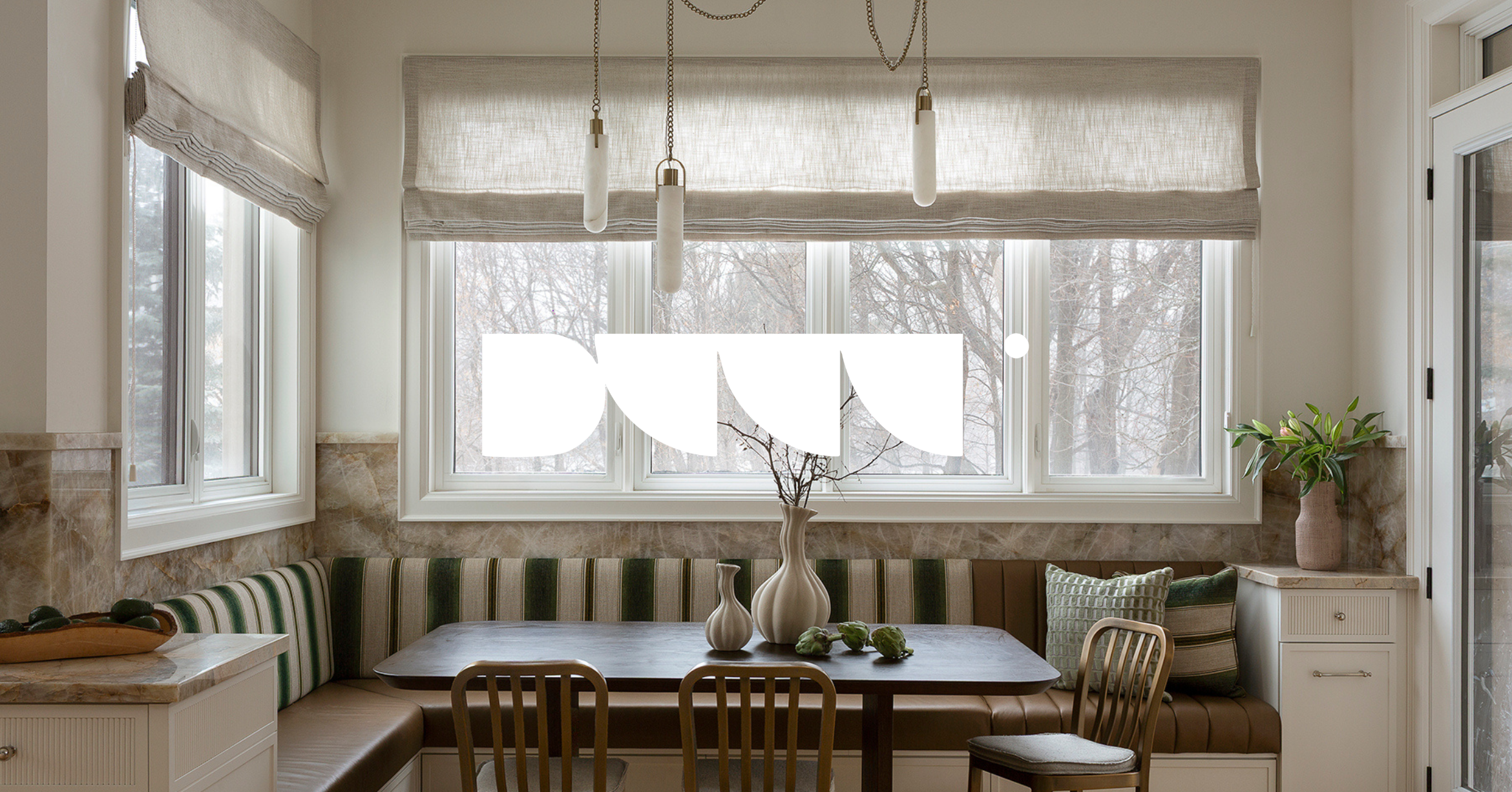 Kitchen nook with large windows covered by beige Roman shades, a table with decorative vases and artichokes, and a padded bench with striped cushions, a bar stool, and a small cabinet with a potted plant.
