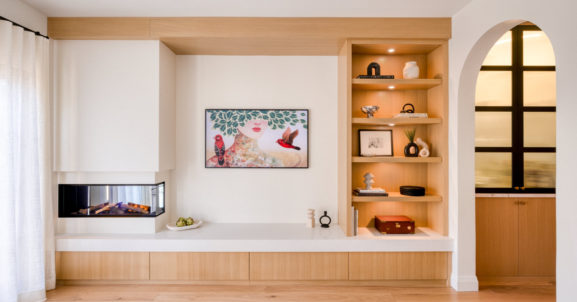 Modern living room with a white fireplace, colorful artwork of a woman with birds, wooden built-in shelves with decorative objects, and a window with black trim.