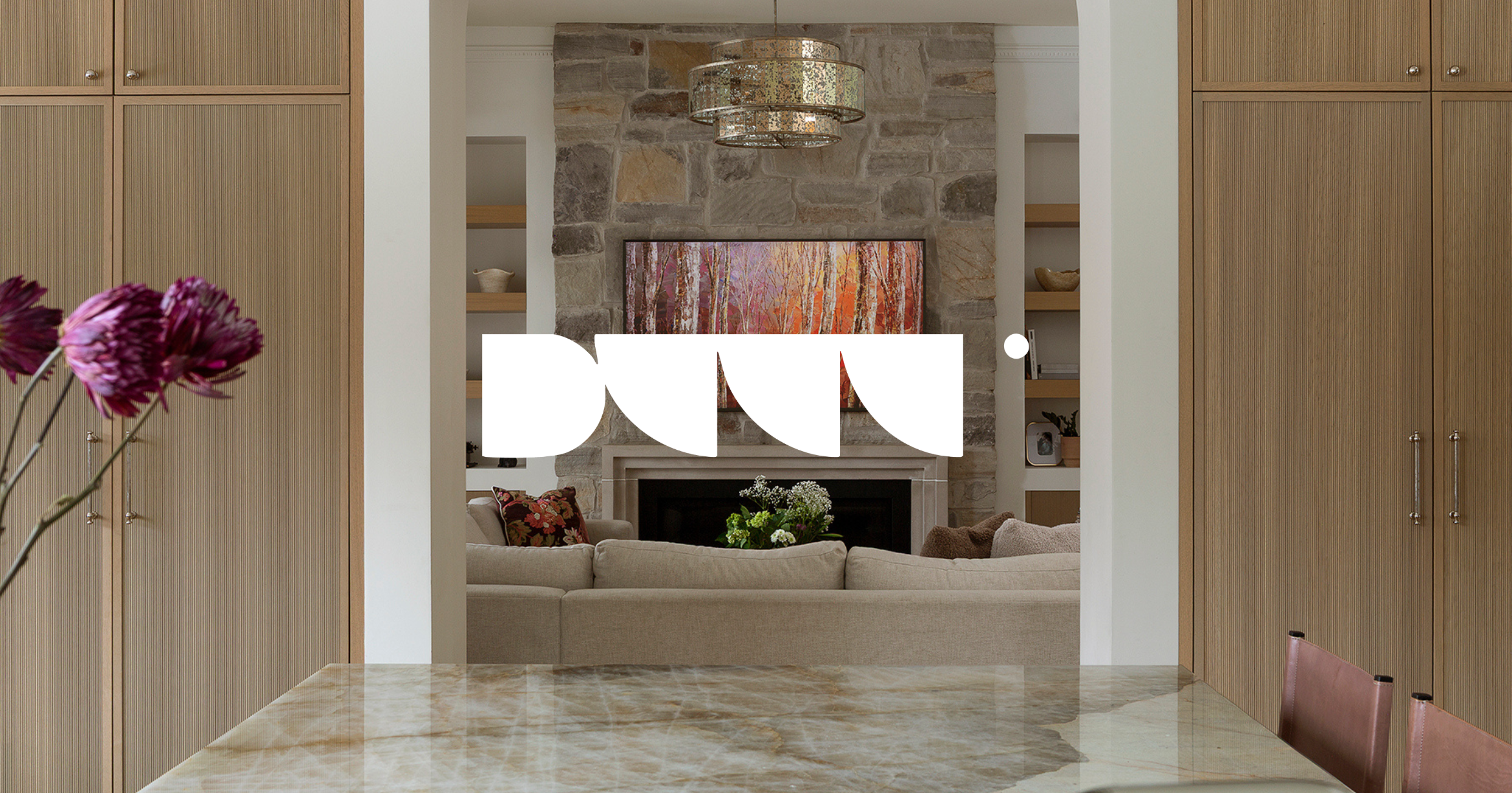 A living room with a stone fireplace, a mounted TV above the fireplace displaying a forest scene, beige sofas, and built-in bookshelves with decorative items. The foreground shows a table with purple flowers.