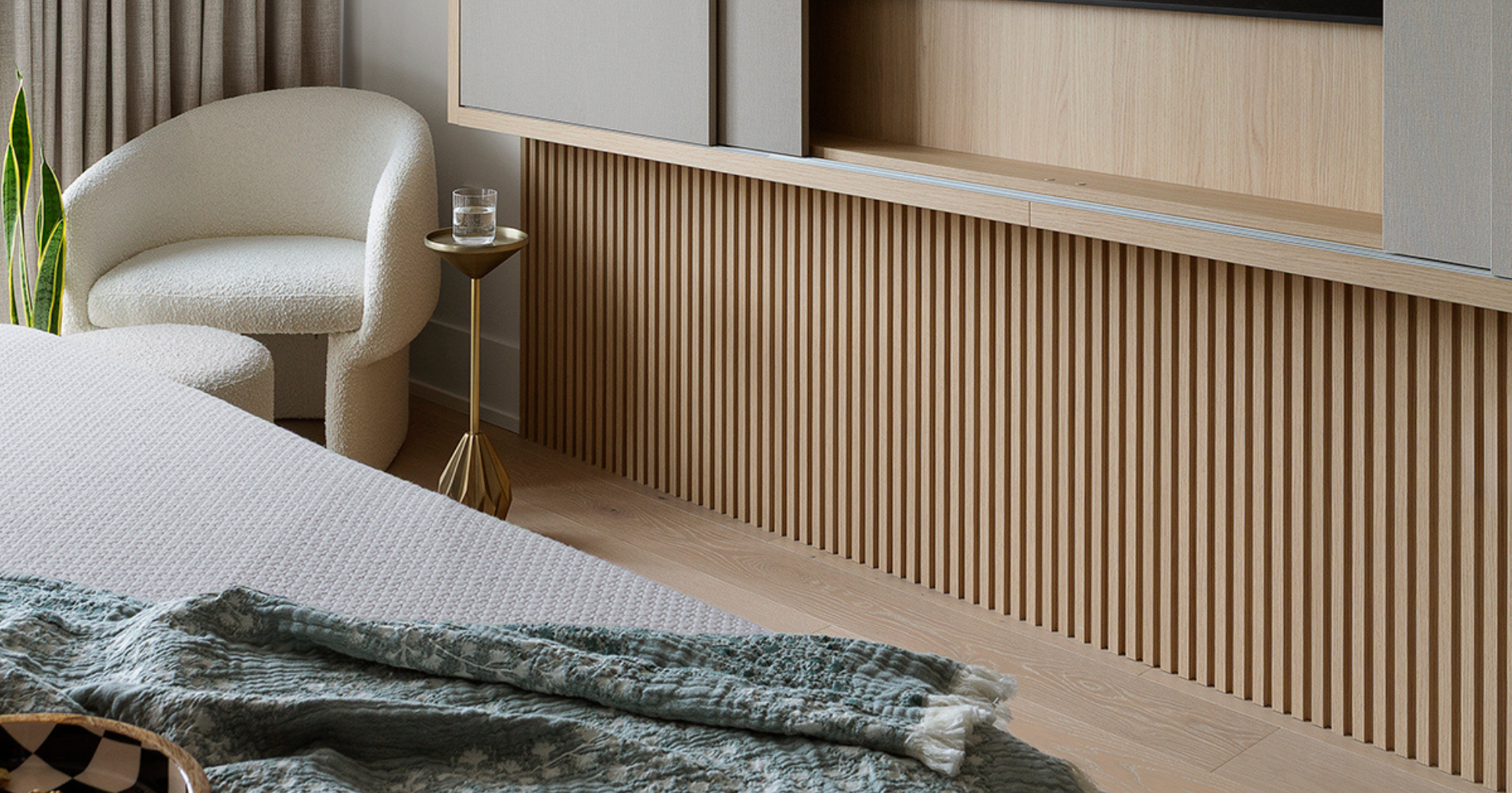 Modern bedroom corner with a white plush armchair, a small gold side table with a glass of water, a bed with a textured blanket, and a wooden slatted cabinet with floating shelves above.