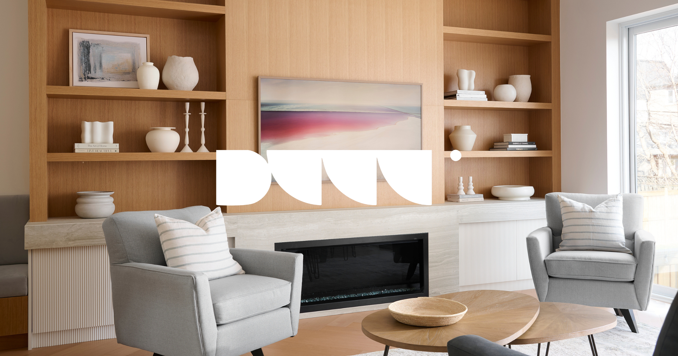Living room with wooden shelving, abstract artwork above a fireplace, two light gray armchairs with striped pillows, a wooden coffee table, and a large window letting in natural light.