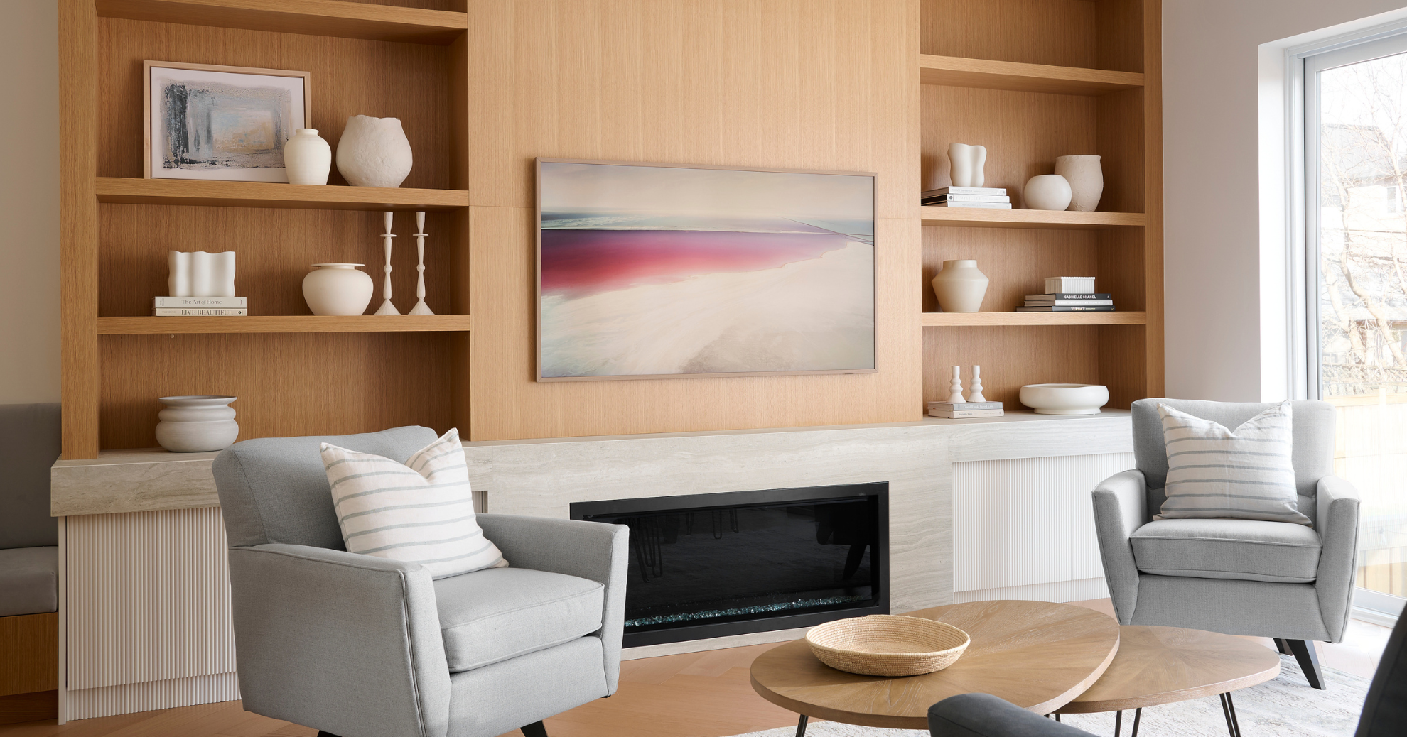 Living room with a built-in wooden shelving unit, modern fireplace, and two gray armchairs with striped pillows. There is a wooden coffee table in front and a large window on the right side.