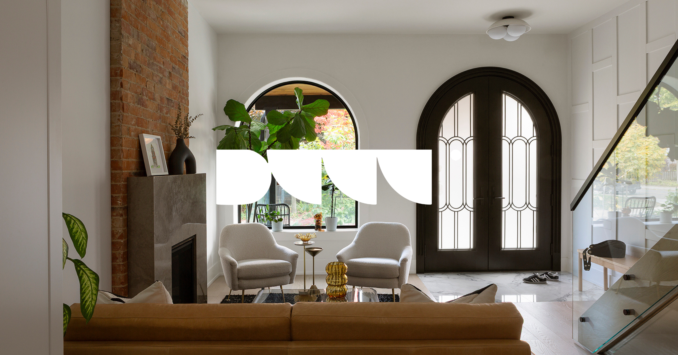 A cozy living room with a white armchair, a small side table, and a beige sofa. There's a large arched window with greenery outside, a dark double-door with decorative glass, and a brick fireplace. Indoor plants and decorative items are also visible.