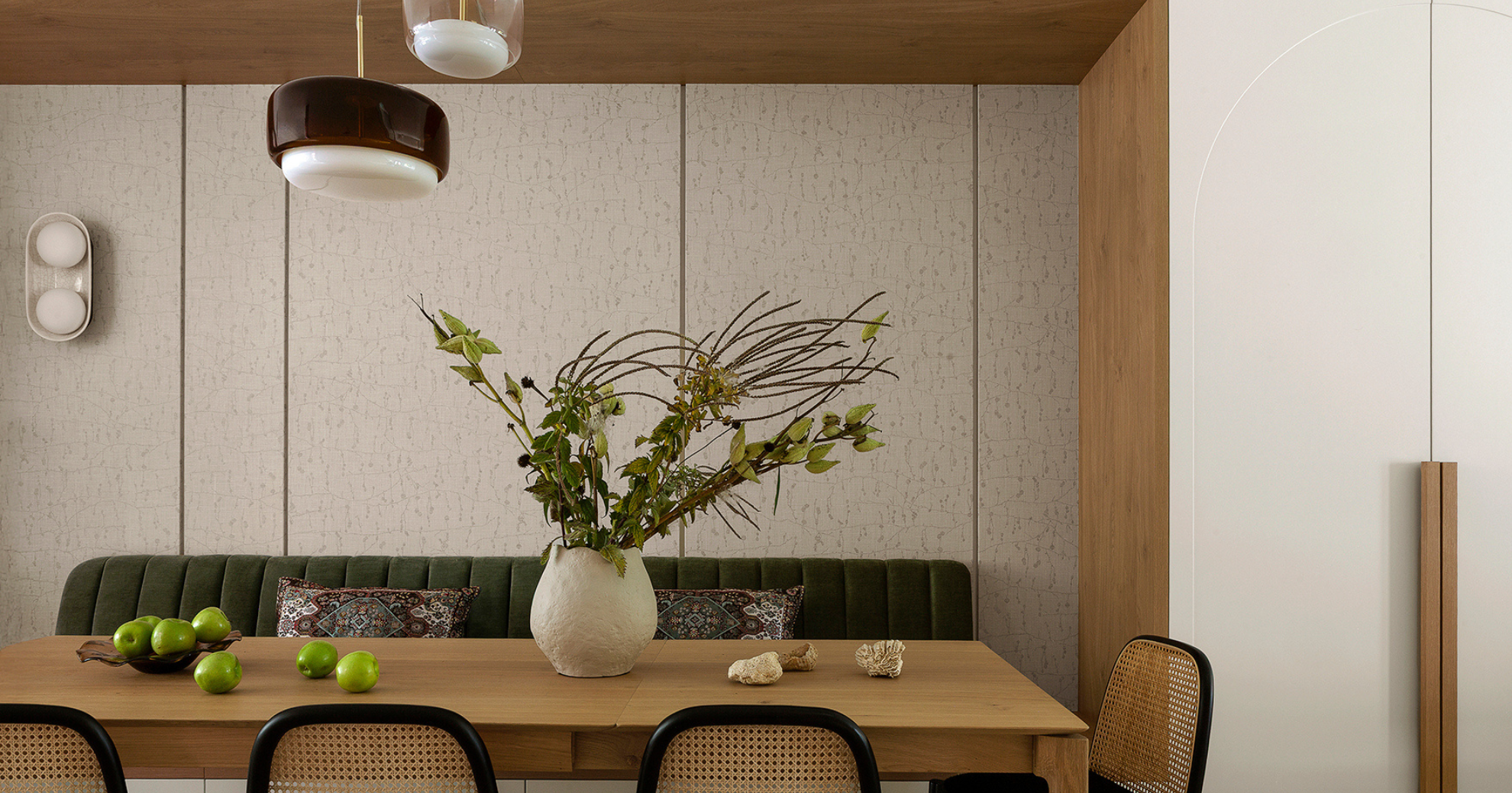 Interior dining room with a wooden table, green patterned bench, rattan chairs, a large white vase with branches, green apples, and decorative shells.