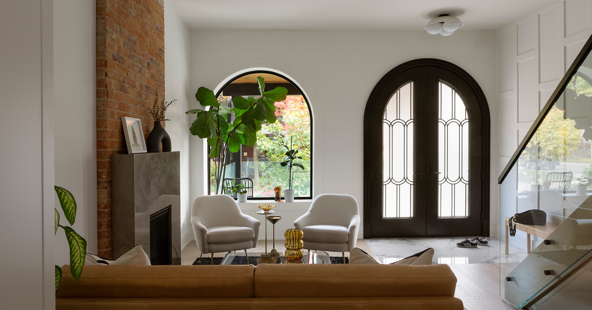 Living room with white walls, a brick fireplace, black double doors, a large green plant, two white armchairs, a small side table, and a window with outdoor view.