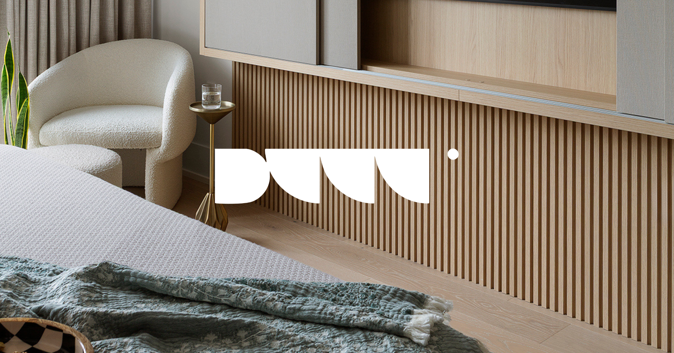 Modern bedroom with a white armchair, a glass of water on a side table, a textured bedspread, and a wooden slatted wall panel.