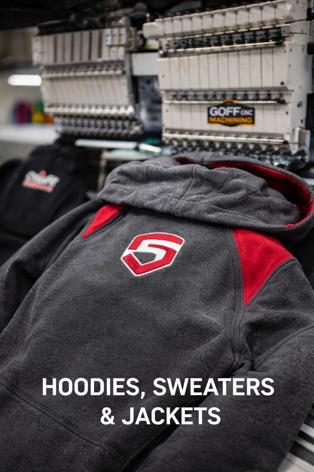 A black and red hoodie with a logo on the chest placed on a surface with other black hoodies or sweatshirts. In the background, there are industrial sewing or embroidery machines and a box with the label "GOFF GNC MACHINING." The image is captioned "HOODIES, SWEATERS & JACKETS."