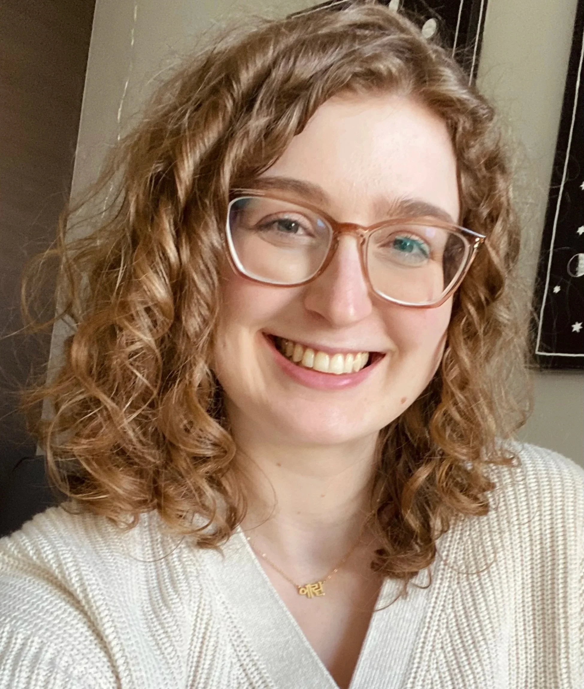 A woman with curly red hair and glasses smiling. She is wearing a cream-colored sweater and a gold necklace with a name tag.