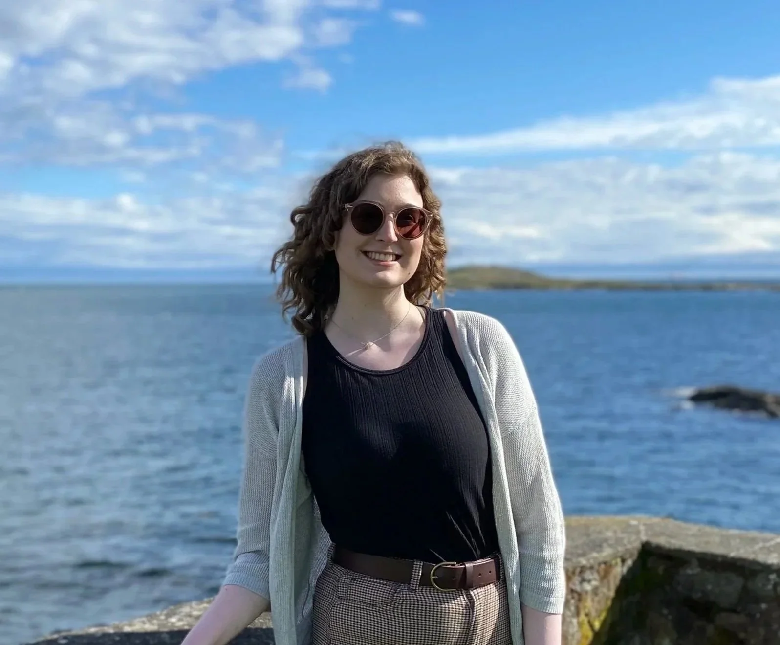 A woman with curly hair and sunglasses smiling at the camera, standing outdoors near a body of water with a cloudy sky in the background.