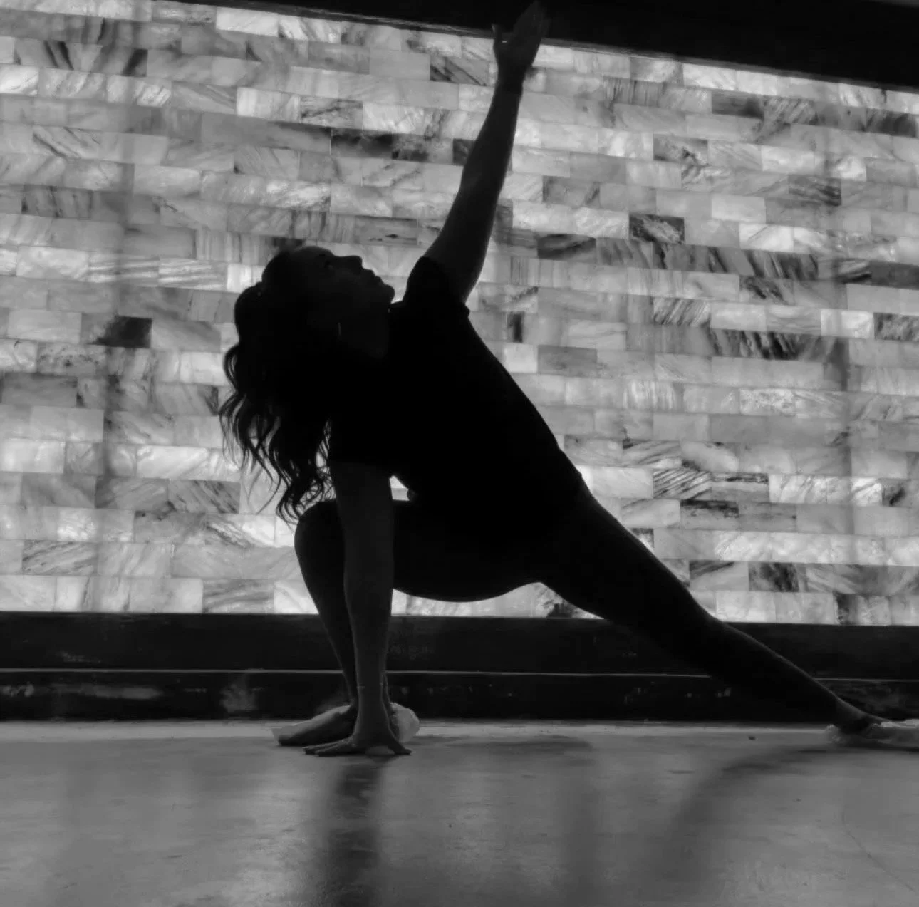 A woman in a yoga pose, silhouetted against a textured wall, with one arm reaching upward and the other supporting her on the floor.