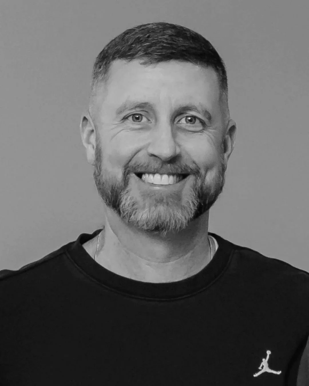 Black and white photo of a smiling man with a beard and short hair, wearing a dark T-shirt with a small Jumpman logo.