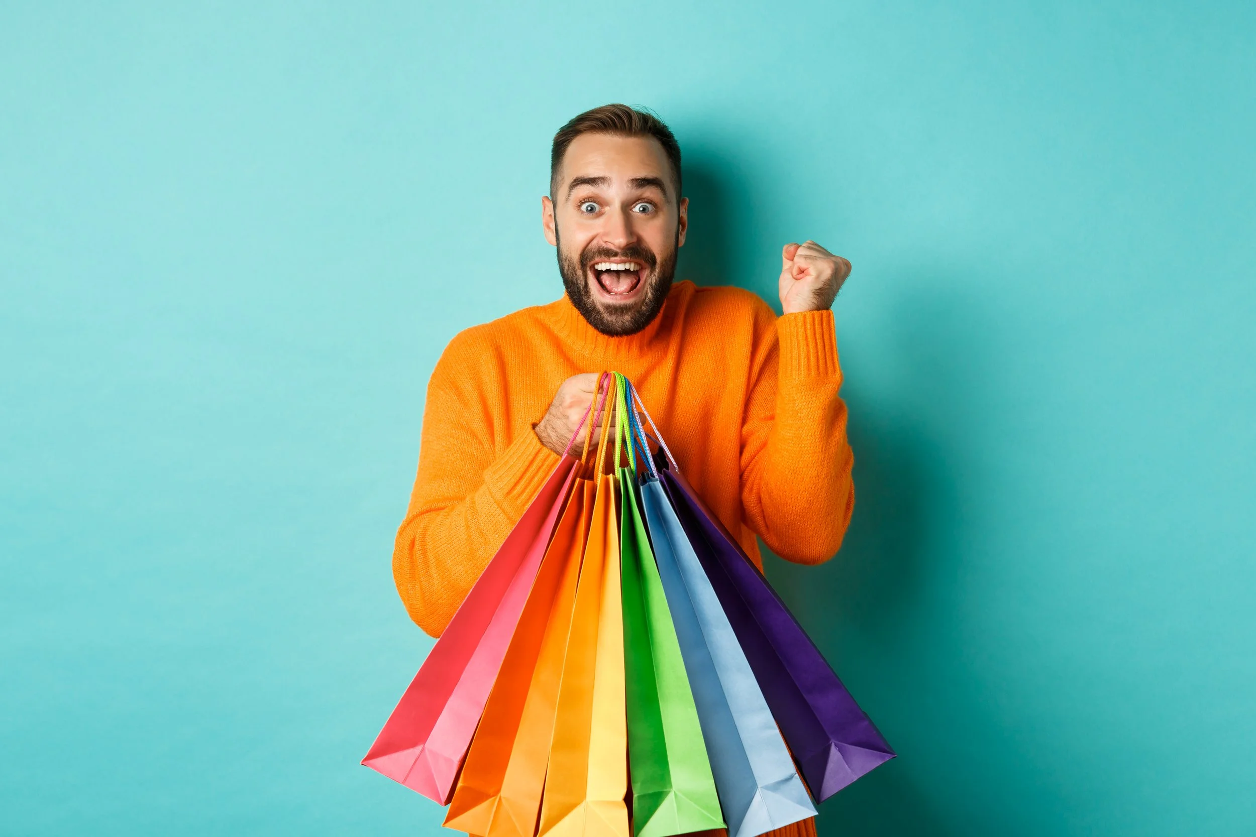 Excited man in orange sweater holding a rainbow array of shopping bags