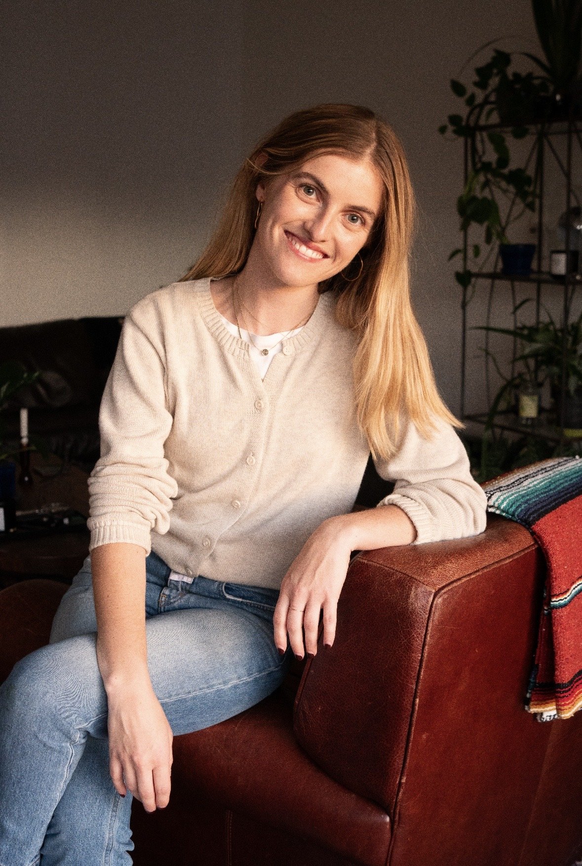A woman with long red hair and blue eyes sitting on a red leather sofa, smiling at the camera, in a cozy living room with plants and a multicolored blanket.