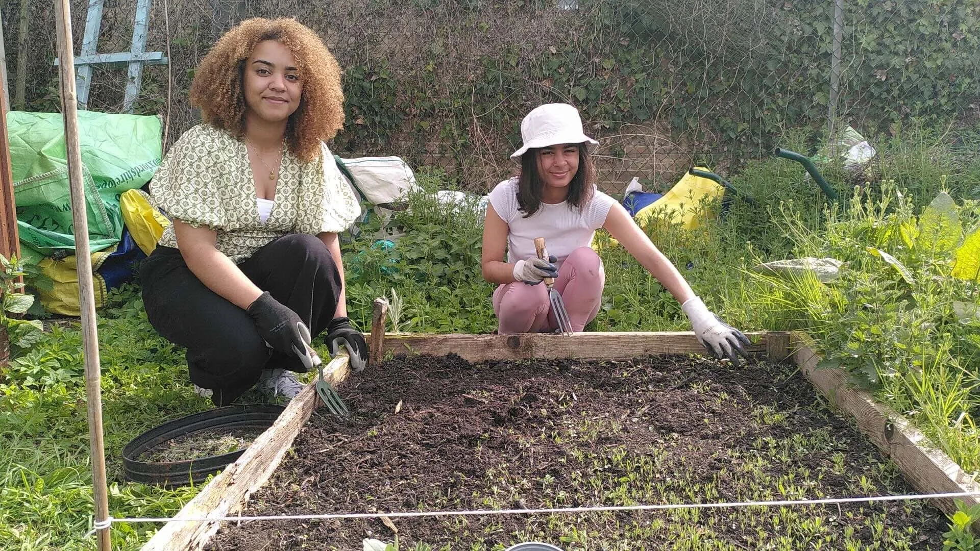 Two women gardening outdoors in an allotment with plants and a metal fence. They are wearing gloves and working on a raised garden bed filled with soil.